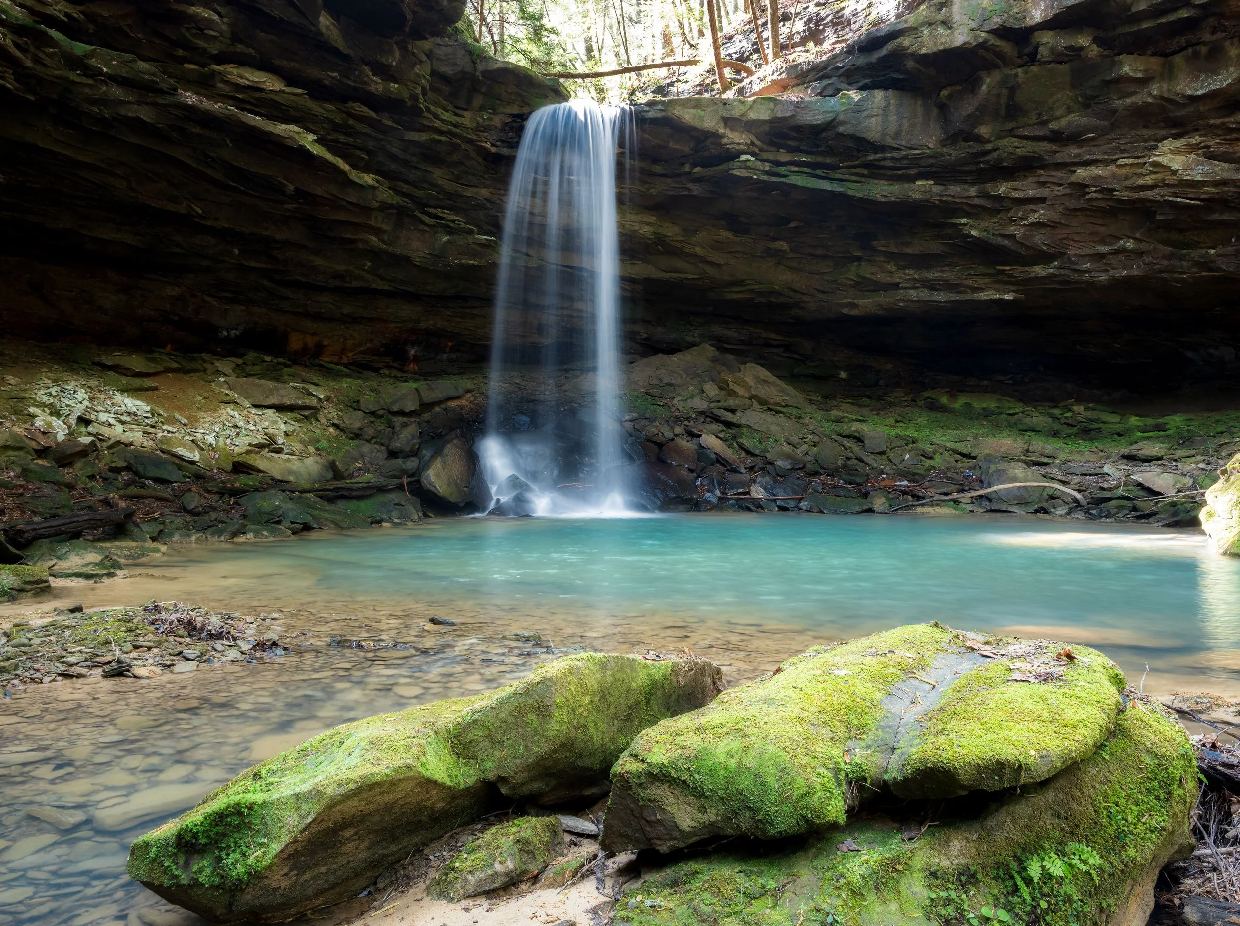 A small waterfall flowing into a clear turquoise pool surrounded by moss-covered rocks and rocks, with a cave-like overhang above and trees in the background.