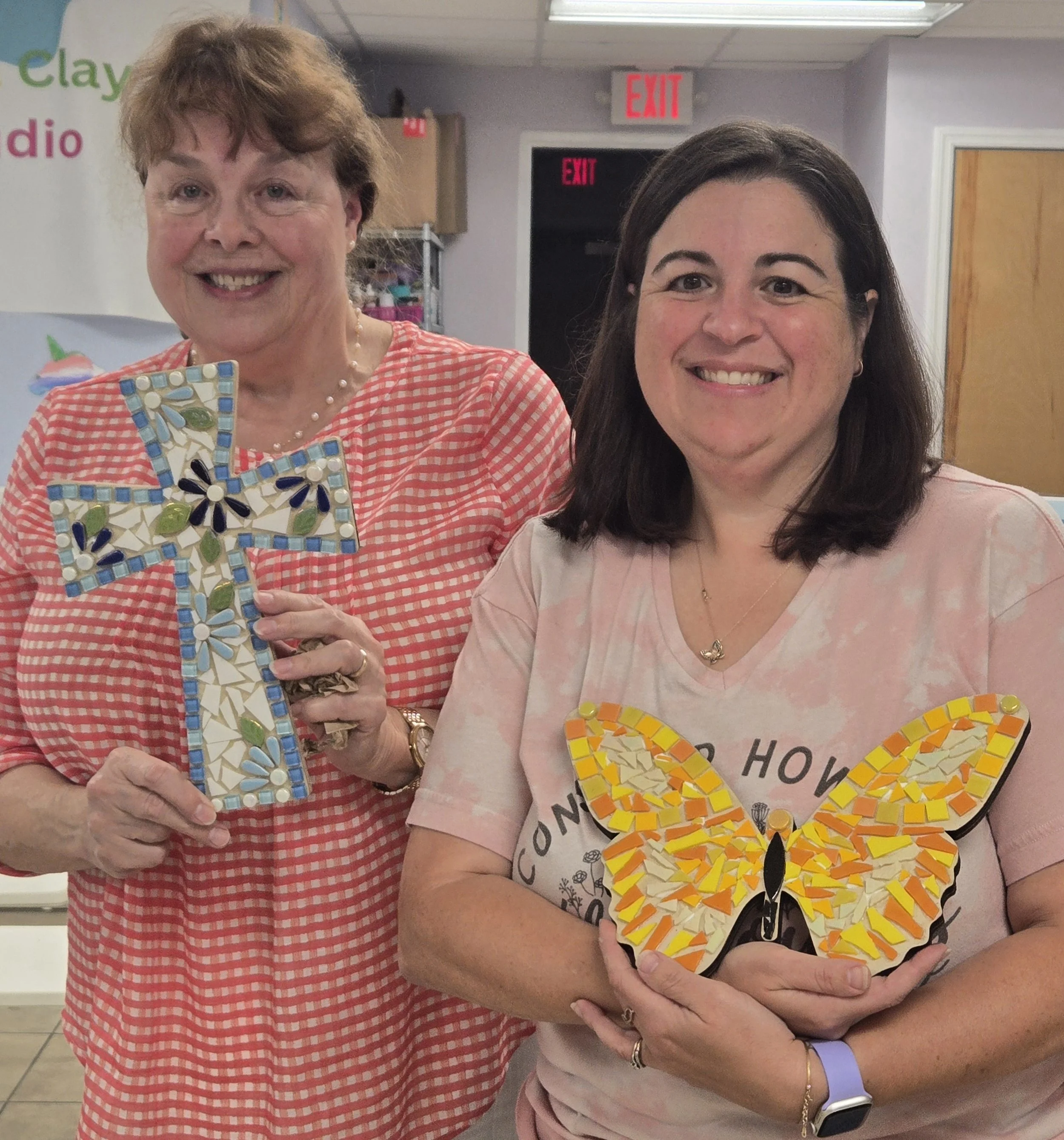 Two women standing close together, smiling, holding colorful mosaic butterfly arts and crafts, in an indoor setting with handmade signs and exit signs in the background.