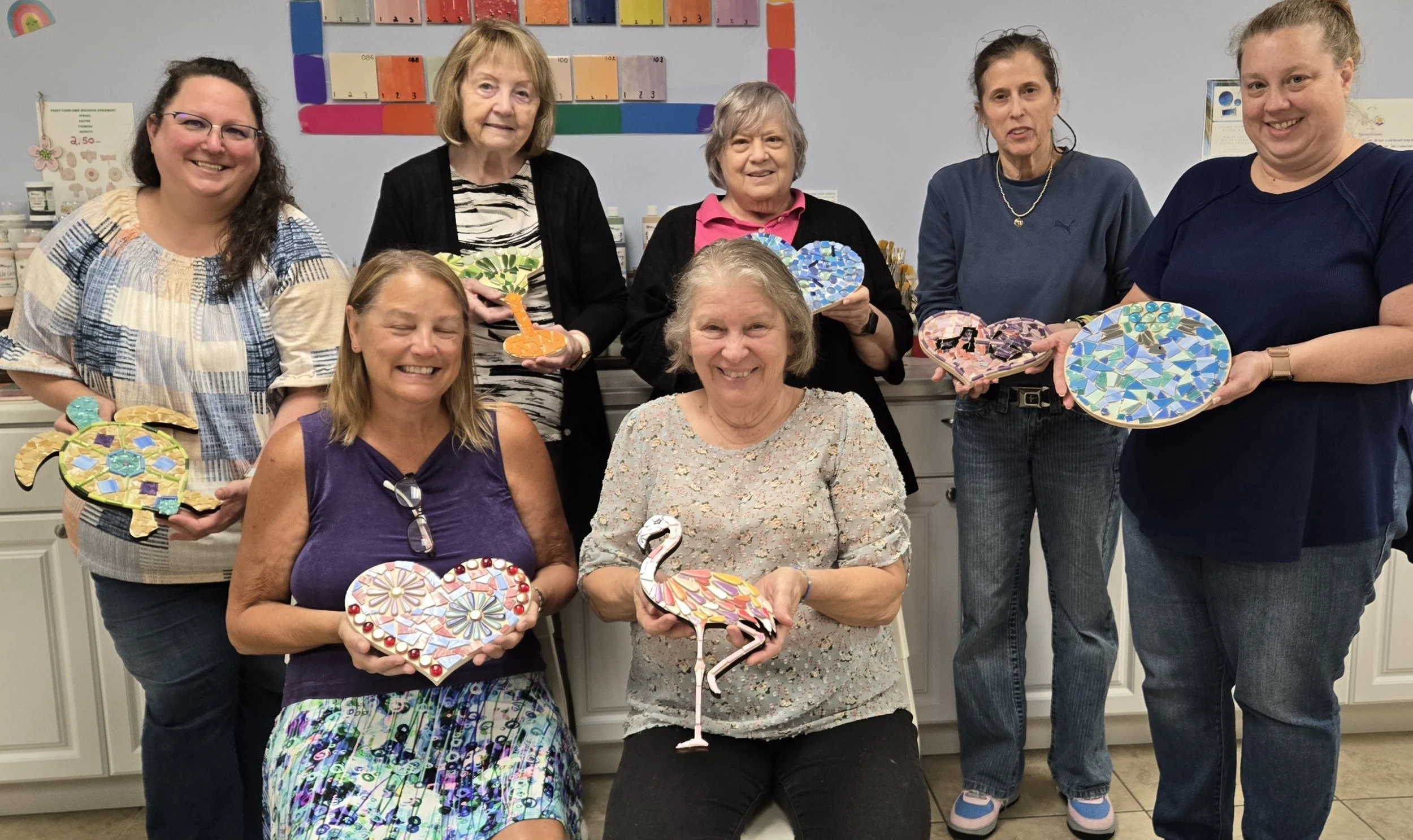 A group of eight women standing and sitting indoors, holding colorful mosaic art pieces, smiling at the camera.