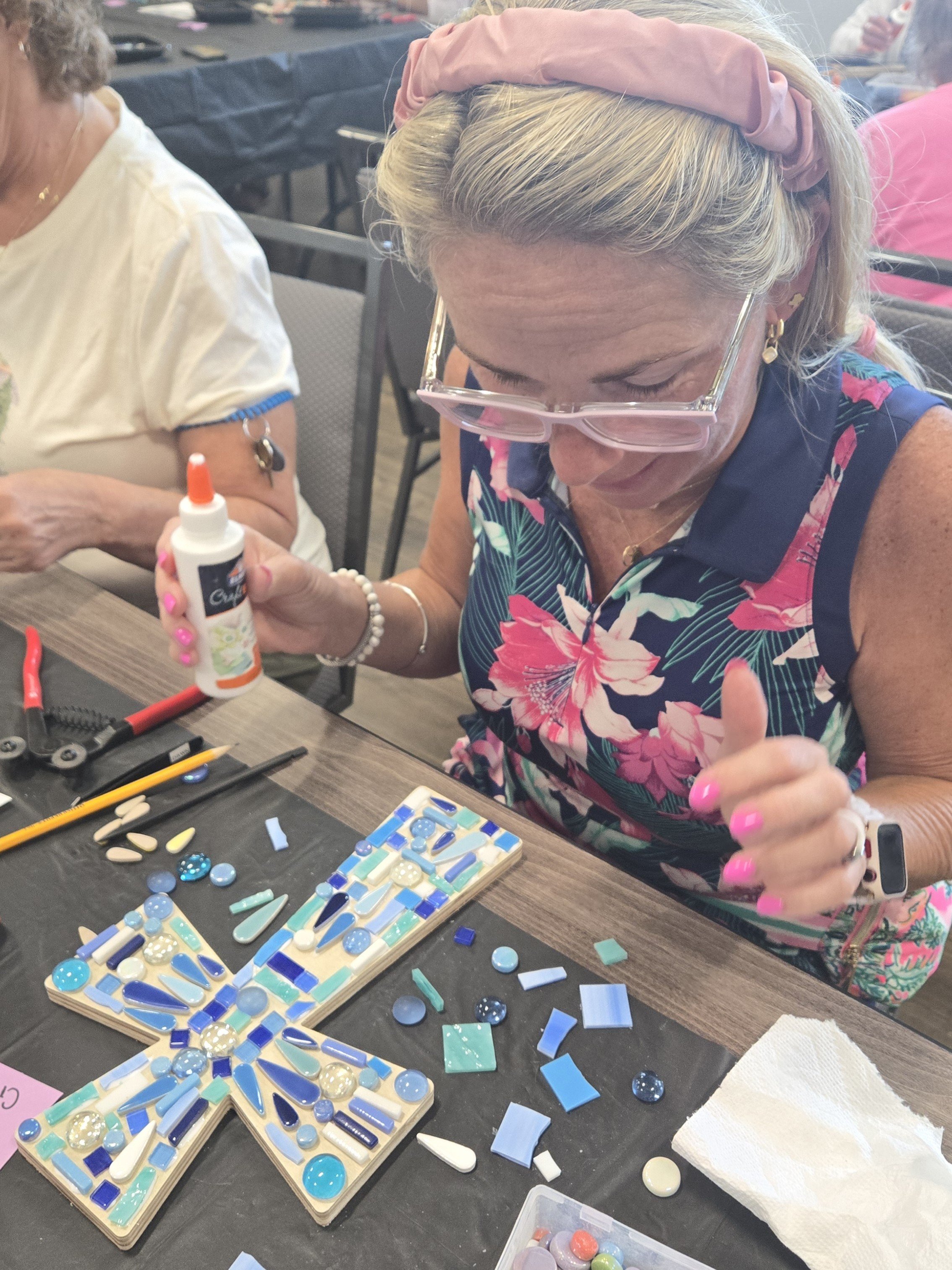 A woman with blonde hair, pink headband, and glasses is working on a mosaic art project using blue, white, and light-colored glass pieces on a wooden board. She is holding a bottle of glue and giving a thumbs-up. There are various mosaic tiles and to