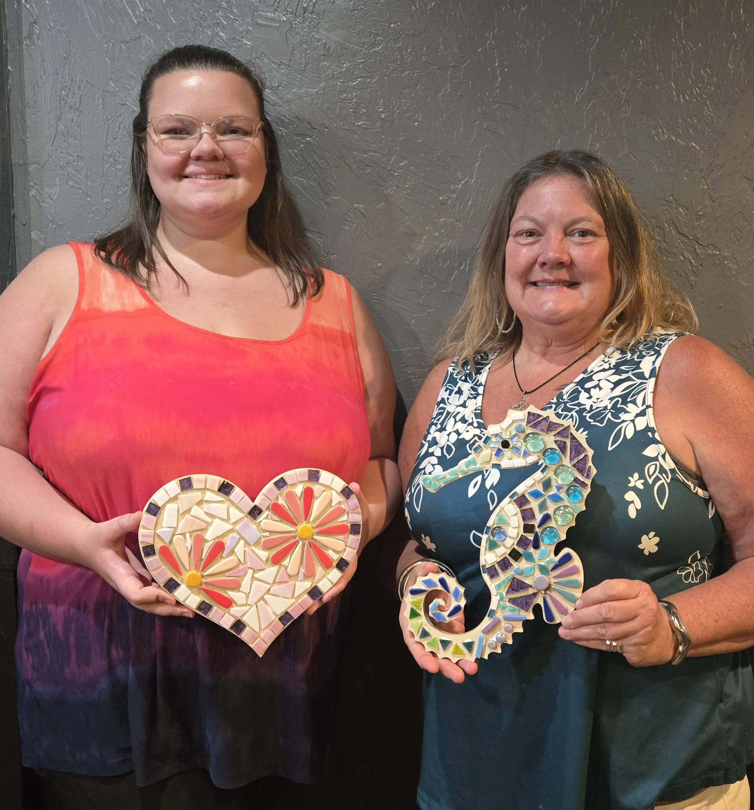 Two women standing against a textured gray wall, holding mosaic heart-shaped and letter-shaped art pieces. The woman on the left wears glasses and an orange sleeveless top, smiling; the woman on the right has long hair, wears a blue and white pattern
