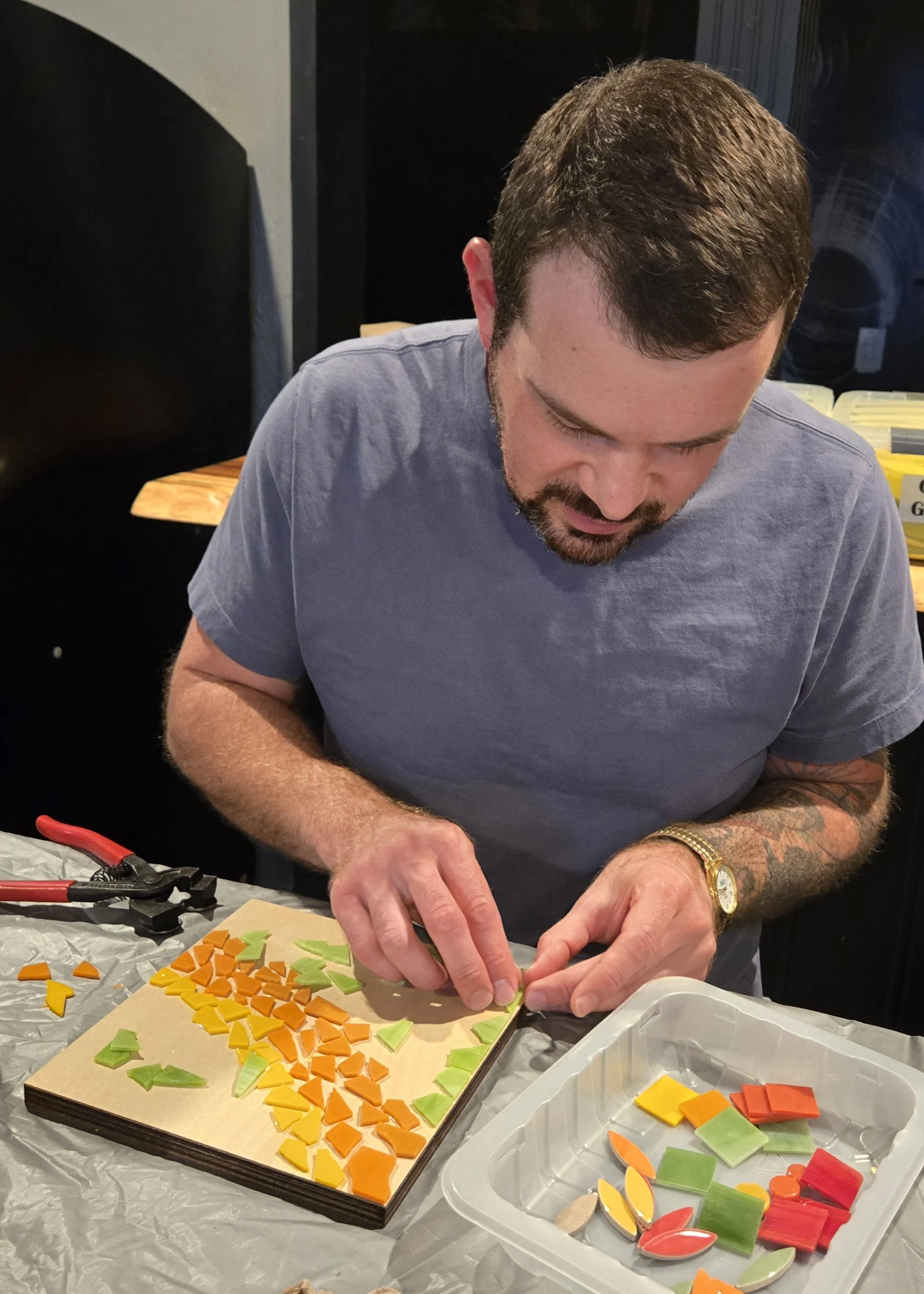 A man with a beard and tattoos is working on mosaic pieces arranged on a tray, with colorful tiles in orange, yellow, green, and red. He is focused on creating a mosaic design, with pliers and a plastic container holding additional tiles nearby.