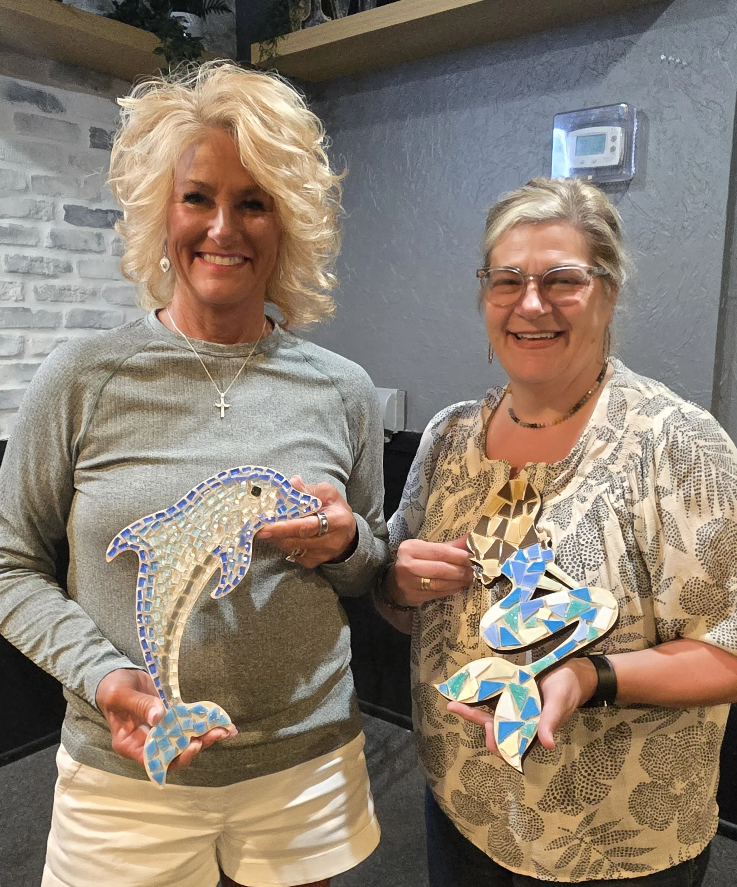 Two women standing side by side, each holding a mosaic art piece of a dolphin. The woman on the left has curly blonde hair, wears a gray sweater and white shorts, and has a necklace with a cross pendant. The woman on the right has straight gray hair,