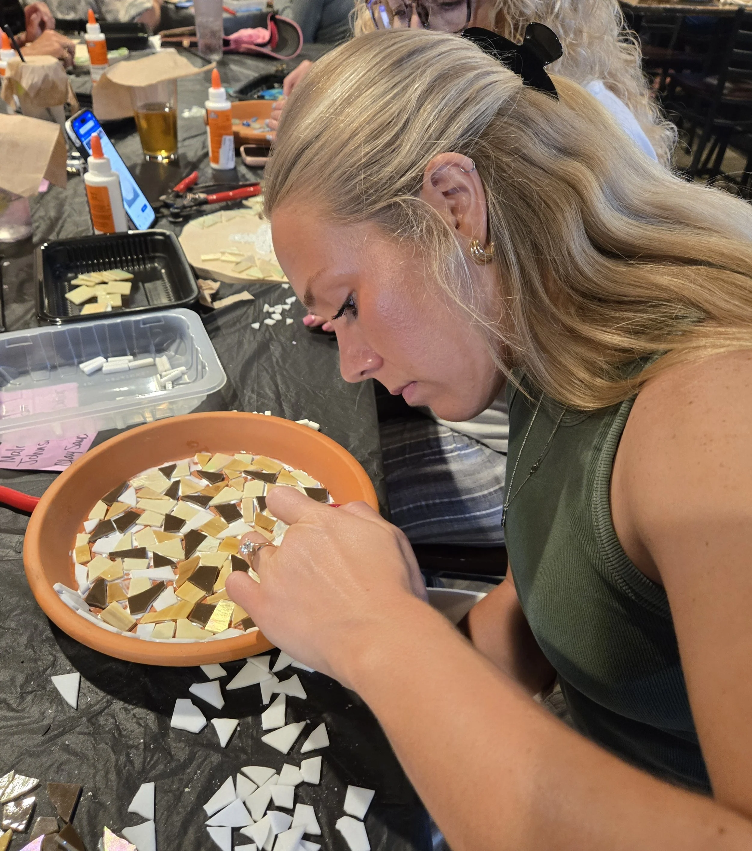 Close-up of a woman with blonde hair and earrings, working on a mosaic art project at a table. She is placing small, reflective, pyramid-shaped tiles into a mosaic design in an orange bowl, on a black table surrounded by art supplies, bottles of glue