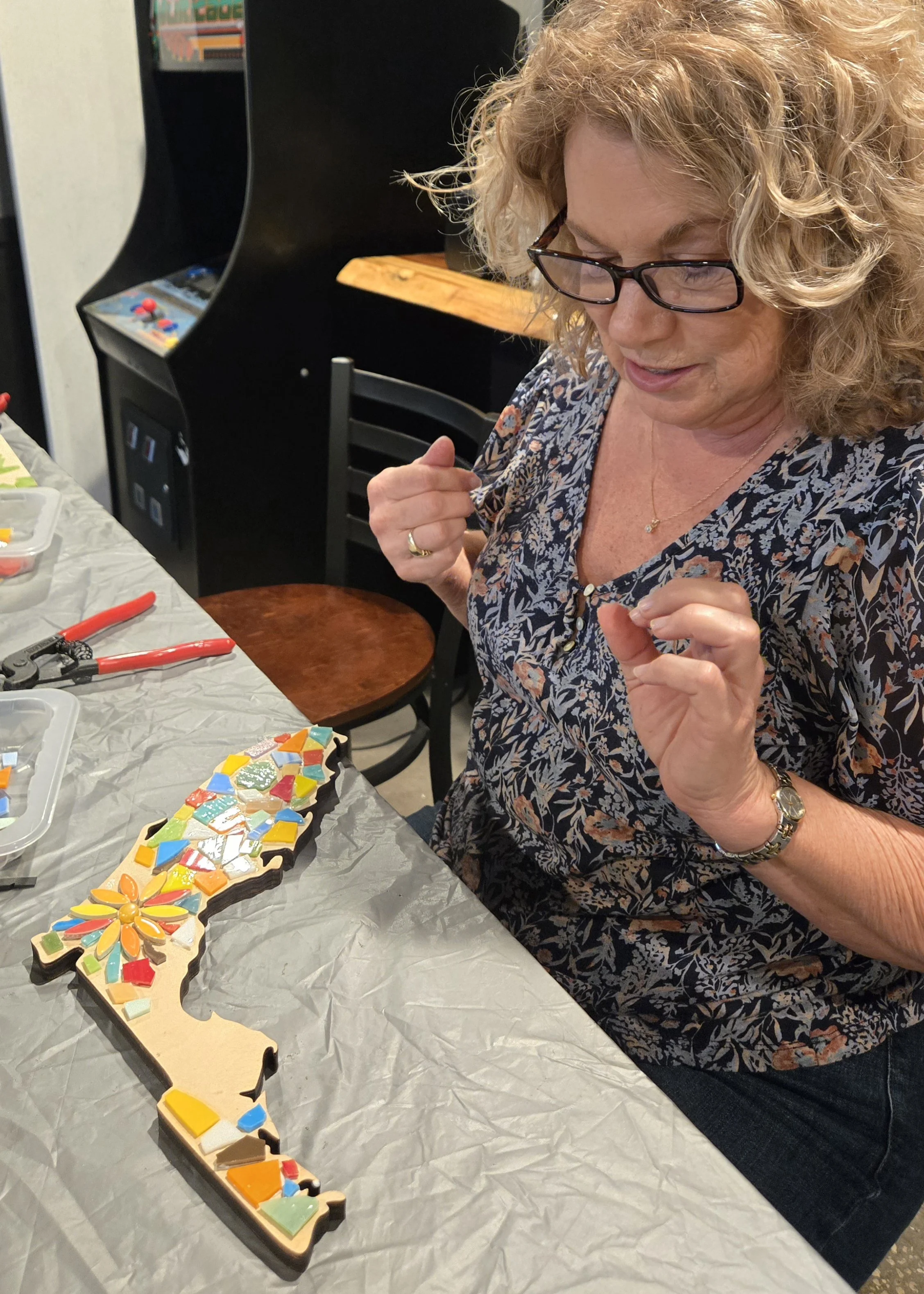 Woman with curly blonde hair and glasses working on a colorful mosaic puzzle at a table covered with a plastic tablecloth. The mosaic pieces are various bright colors, and there are tools like pliers and scissors nearby.