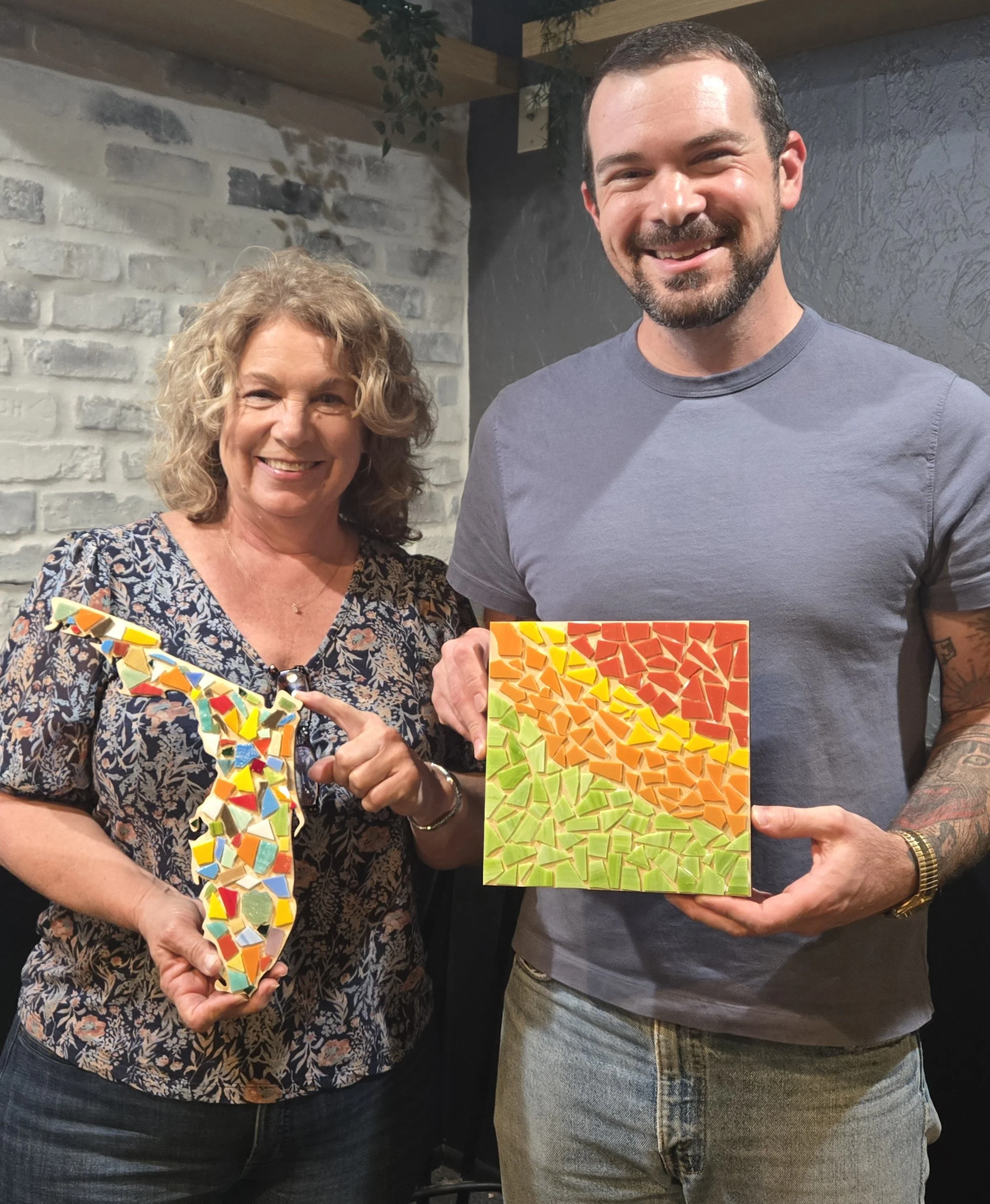 Two people standing side by side, holding colorful mosaic artwork. The woman on the left has curly blonde hair and is wearing a floral blouse. The man on the right has short dark hair, a beard, and is wearing a gray t-shirt and jeans. The background 