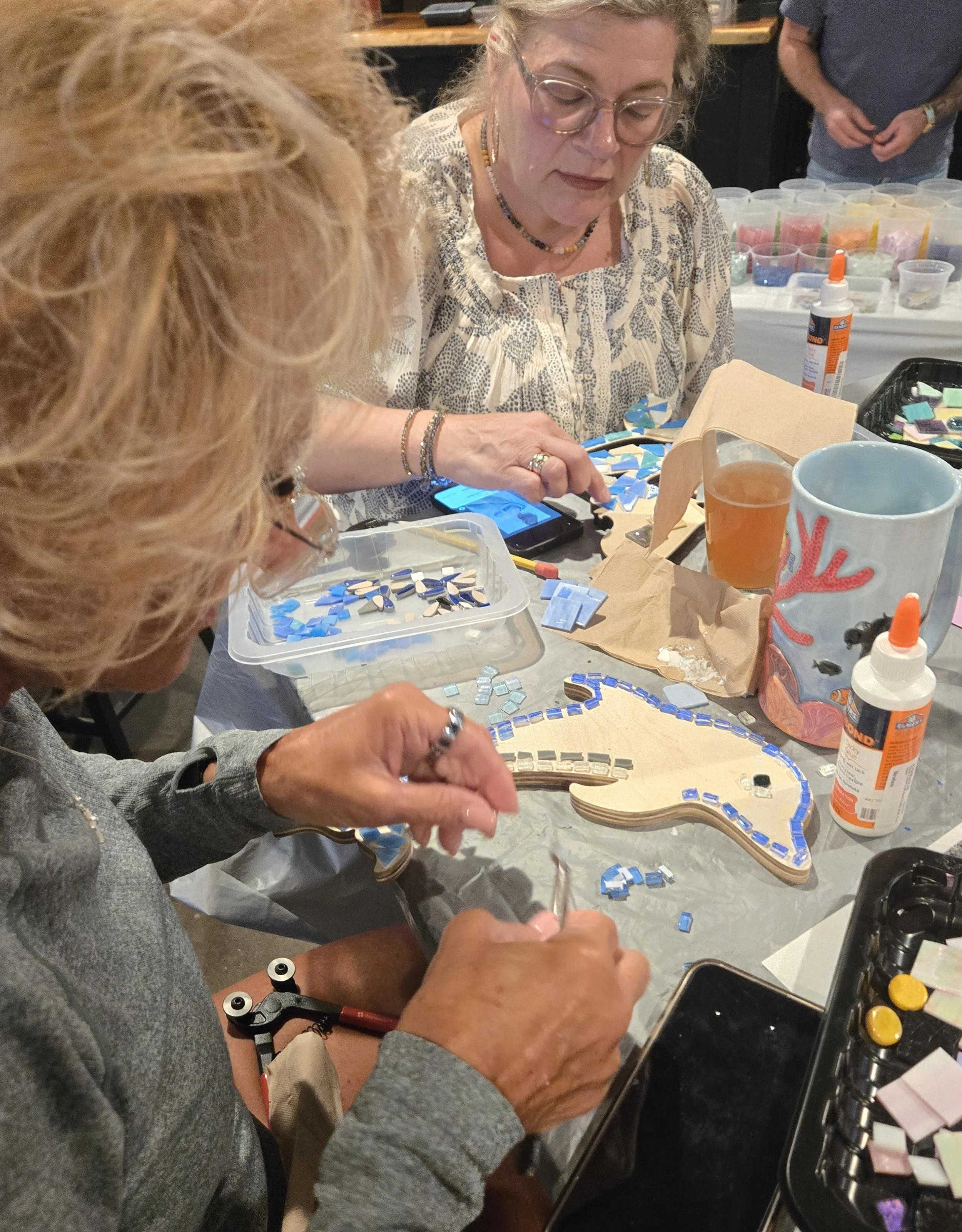 People working on a mosaic craft project with colorful tiles in a busy indoor setting.