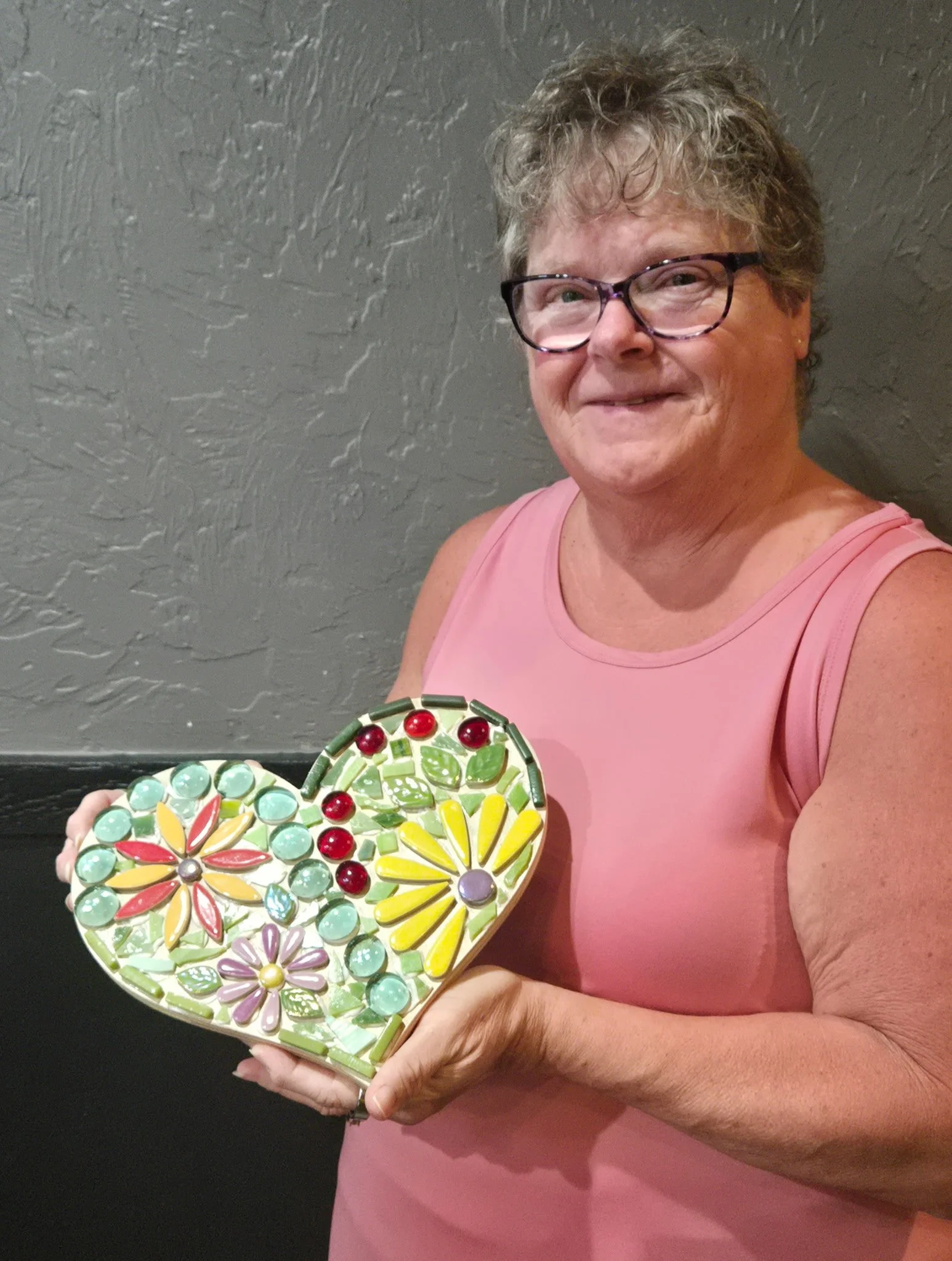 A woman with short curly gray hair wearing glasses and a pink sleeveless top, holding a heart-shaped mosaic art piece with colorful flower designs, standing against a gray textured wall.