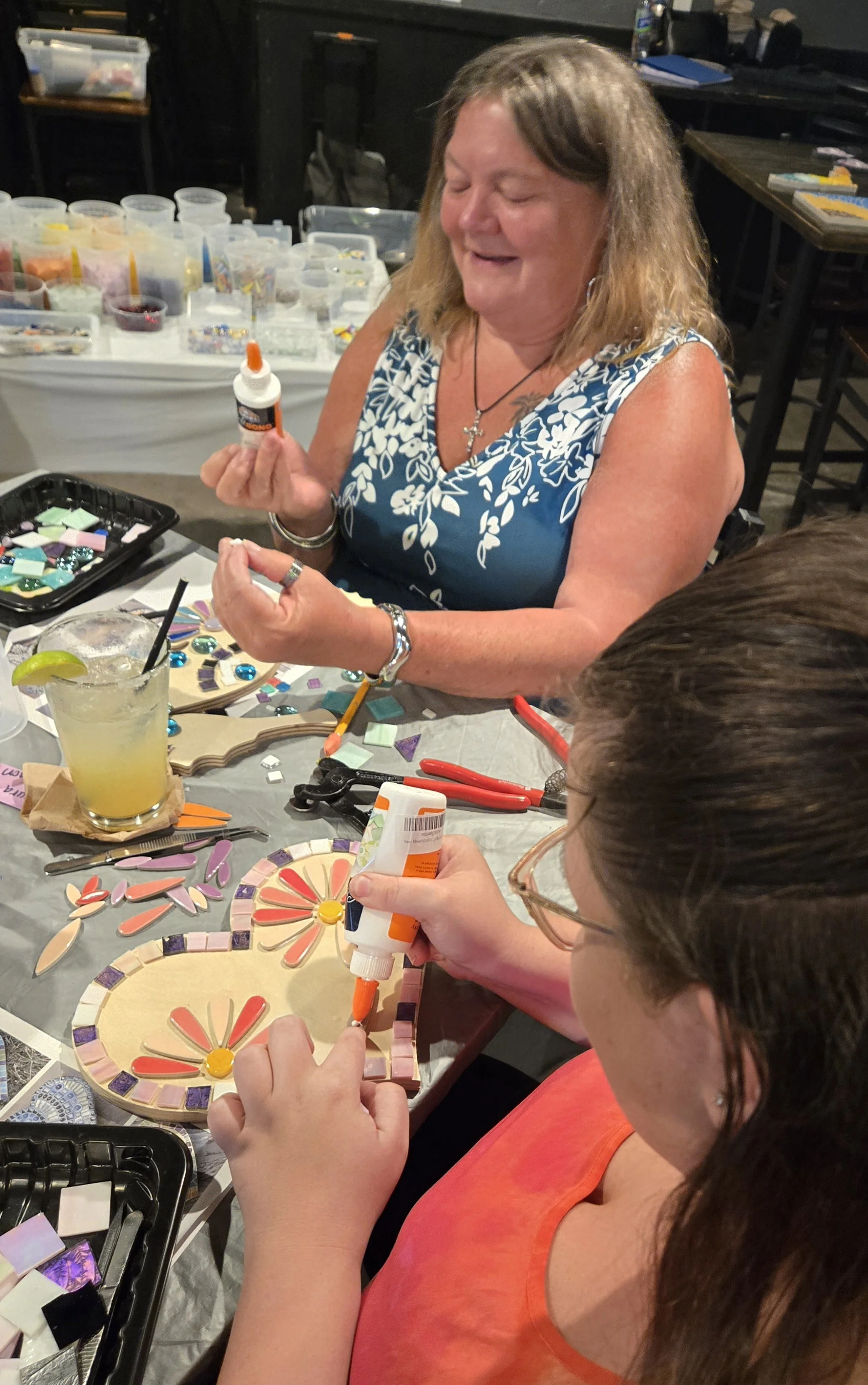 Two women decorating mosaic art pieces at a craft table, surrounded by supplies, glass, and glue bottles.