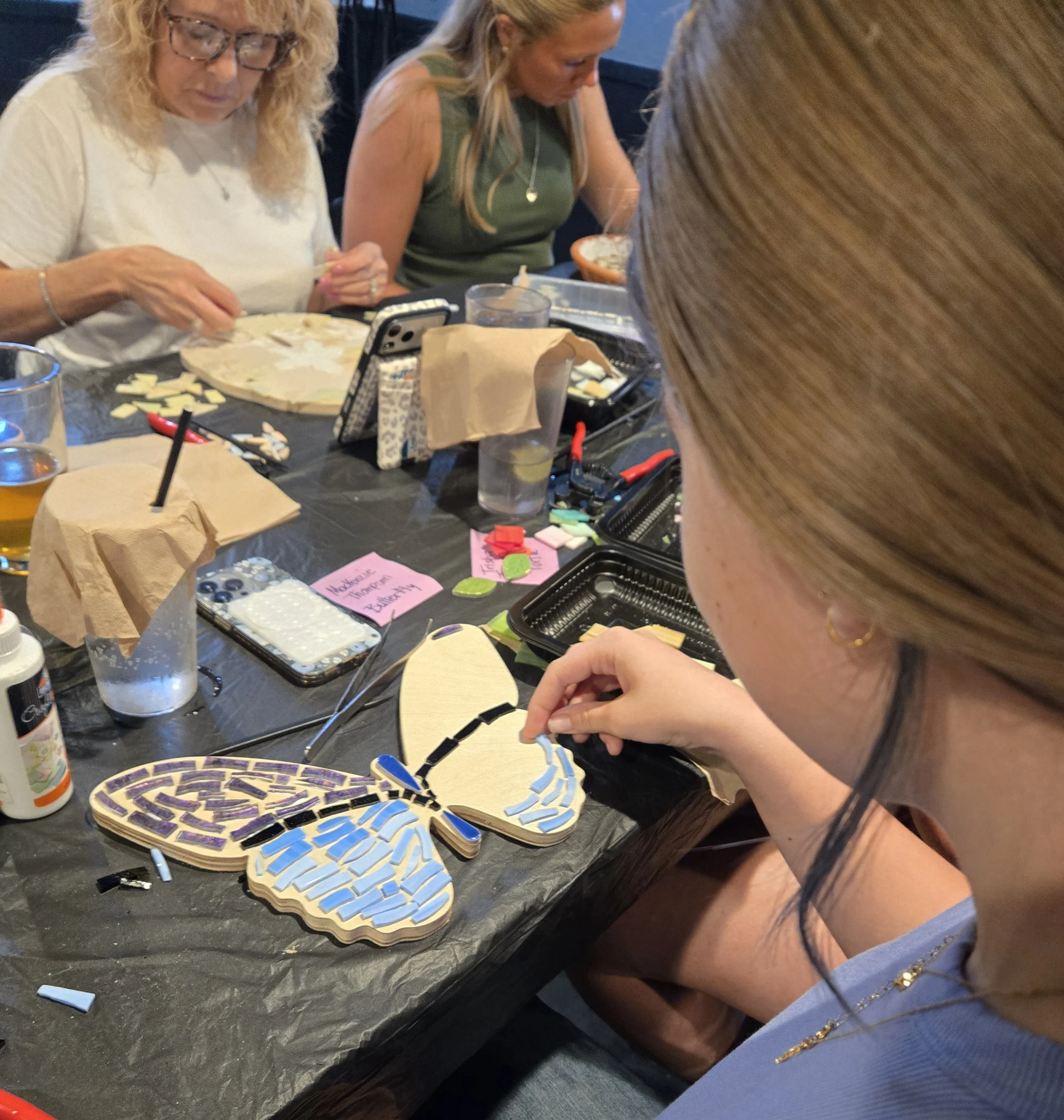 Group of women sitting at a table working on puzzle pieces, with various tools, glue, and notes around them.