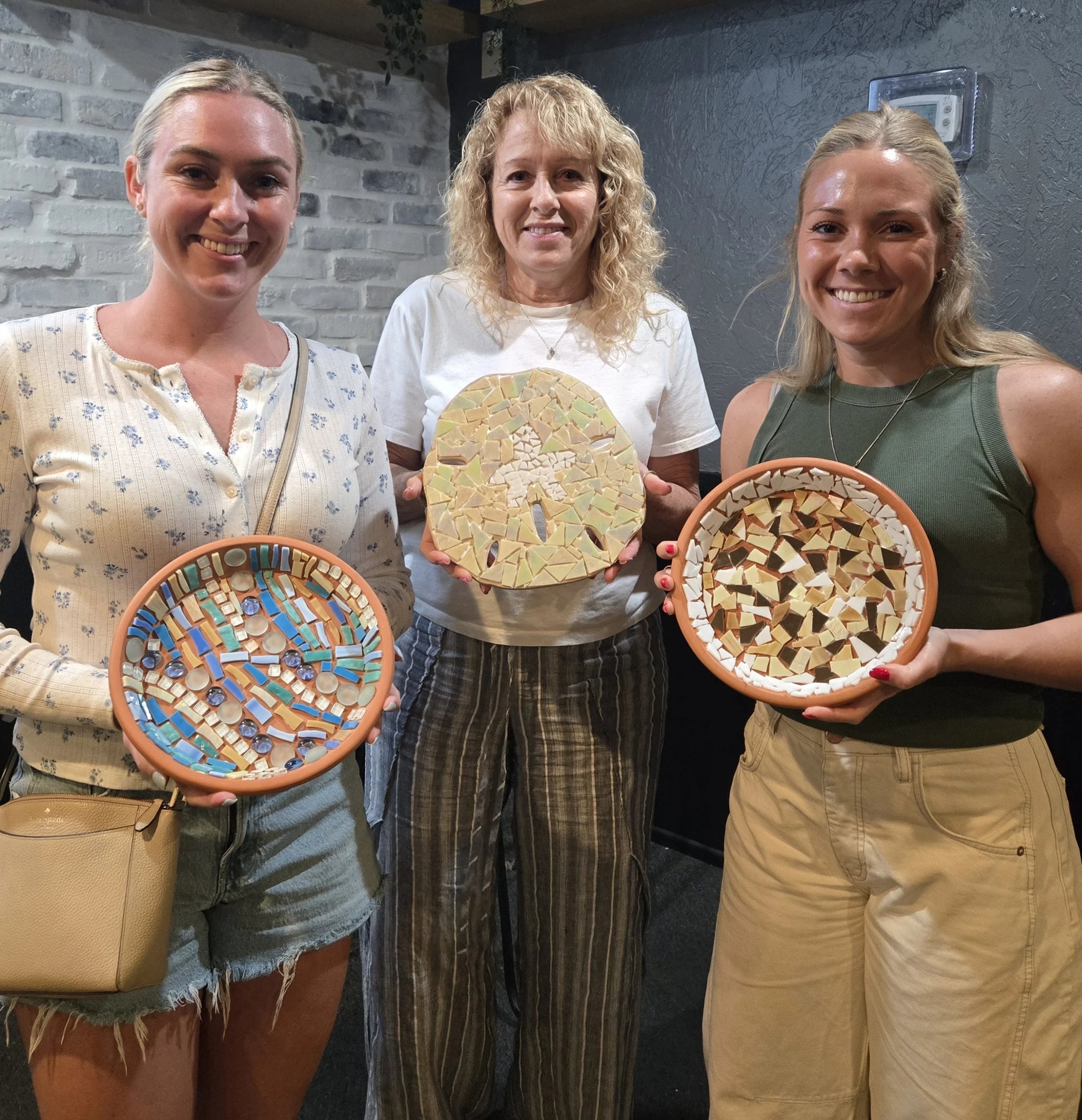 Three women standing side by side indoors, each holding a mosaic art piece with a circular shape. The background includes a gray textured wall and a brick wall, with a small electronic device mounted on the wall to the right.