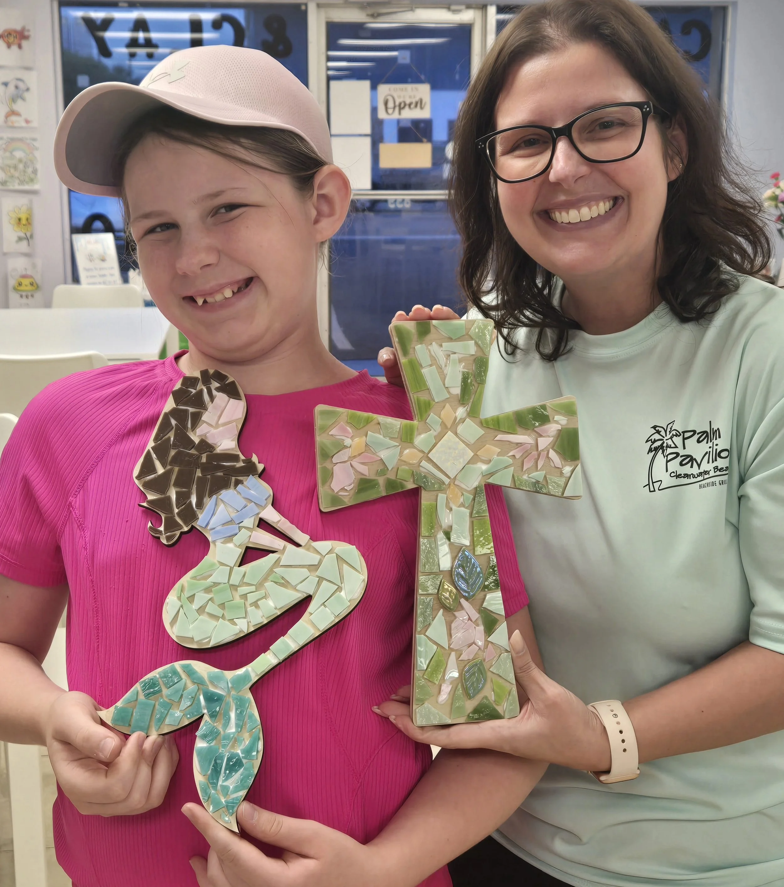 Two women smiling and holding large mosaic dolphin and lighthouse decorations, standing inside a store with glass doors and colorful artwork on the walls.