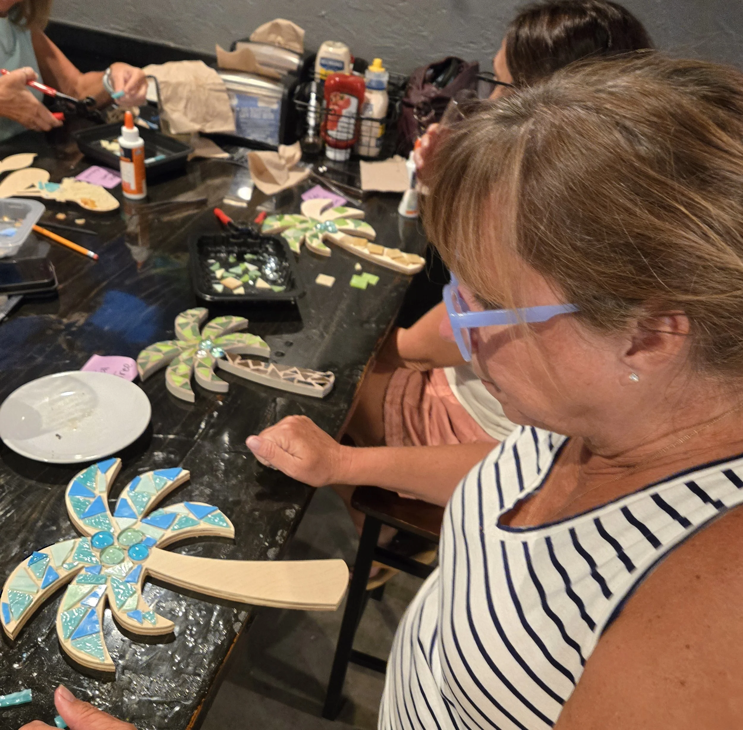 People gathered around a table making mosaic crafts with wooden bases, colorful glass tiles, glue, and scissors.