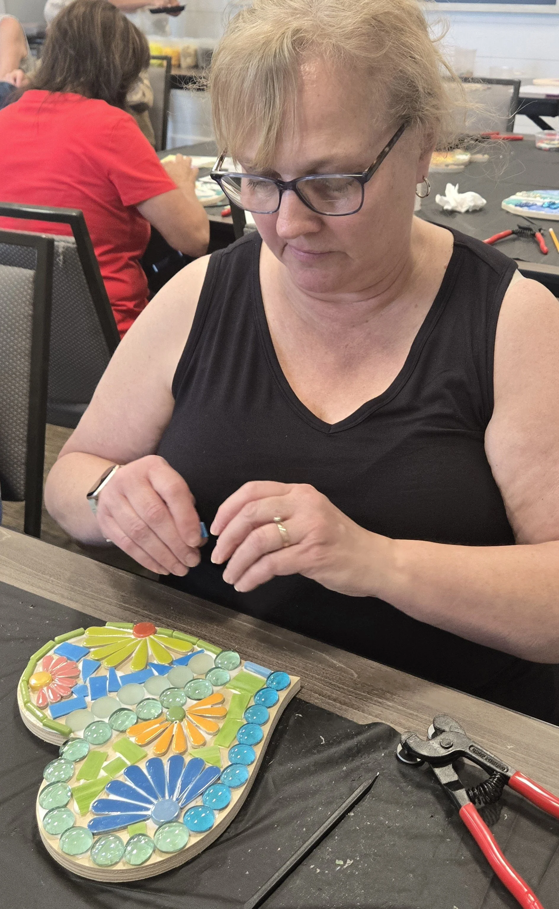 A woman with glasses and hoop earrings sits at a table working on a colorful mosaic art project. The mosaic features flower and petal designs with various colored glass pieces, surrounded by tools like pliers and a glue gun. In the background, anothe