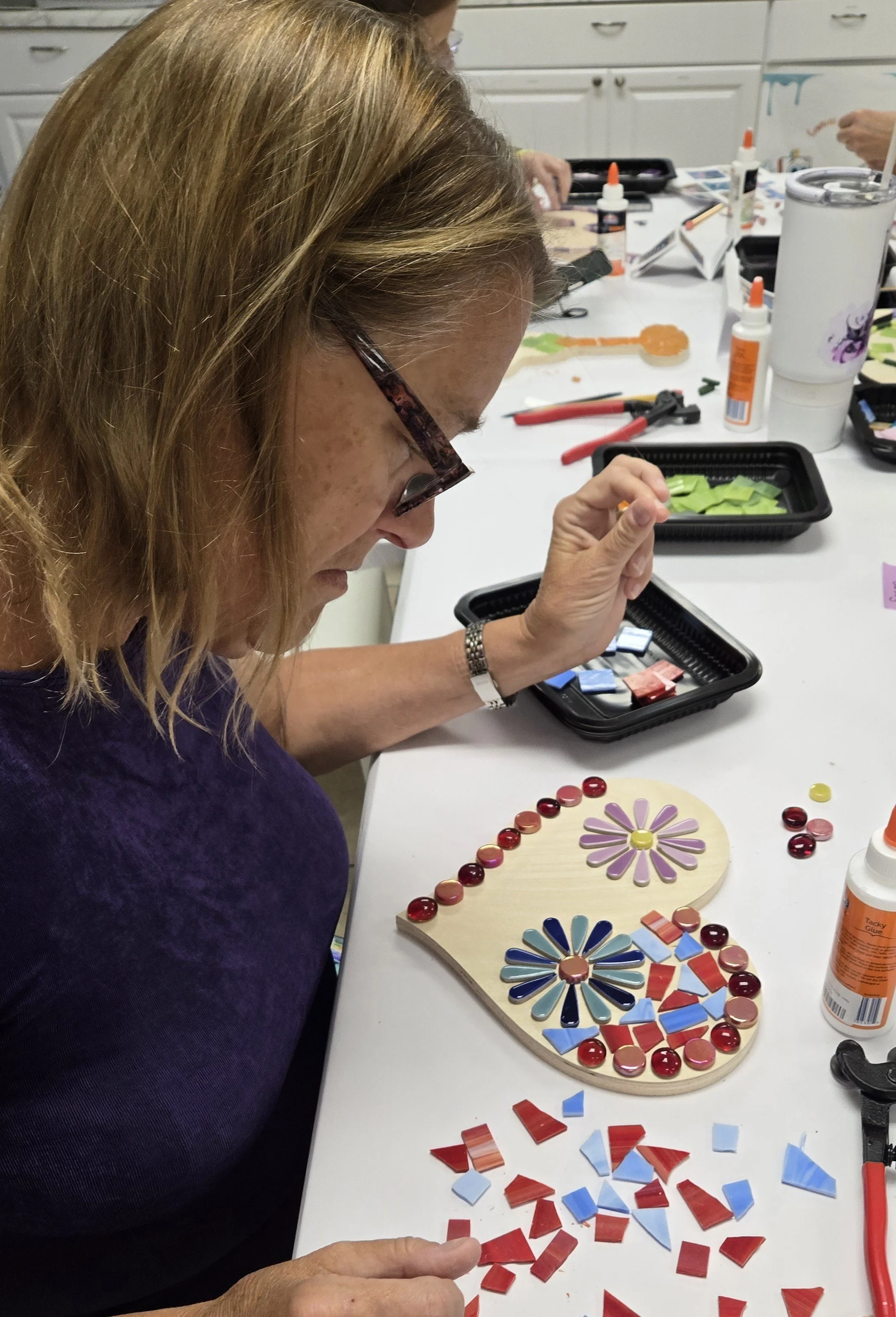 A woman appears to be working on a craft project with colorful mosaic tiles, arranged in a floral pattern on a wooden board, at a table with various crafting supplies like glue bottles, scissors, and additional tiles.