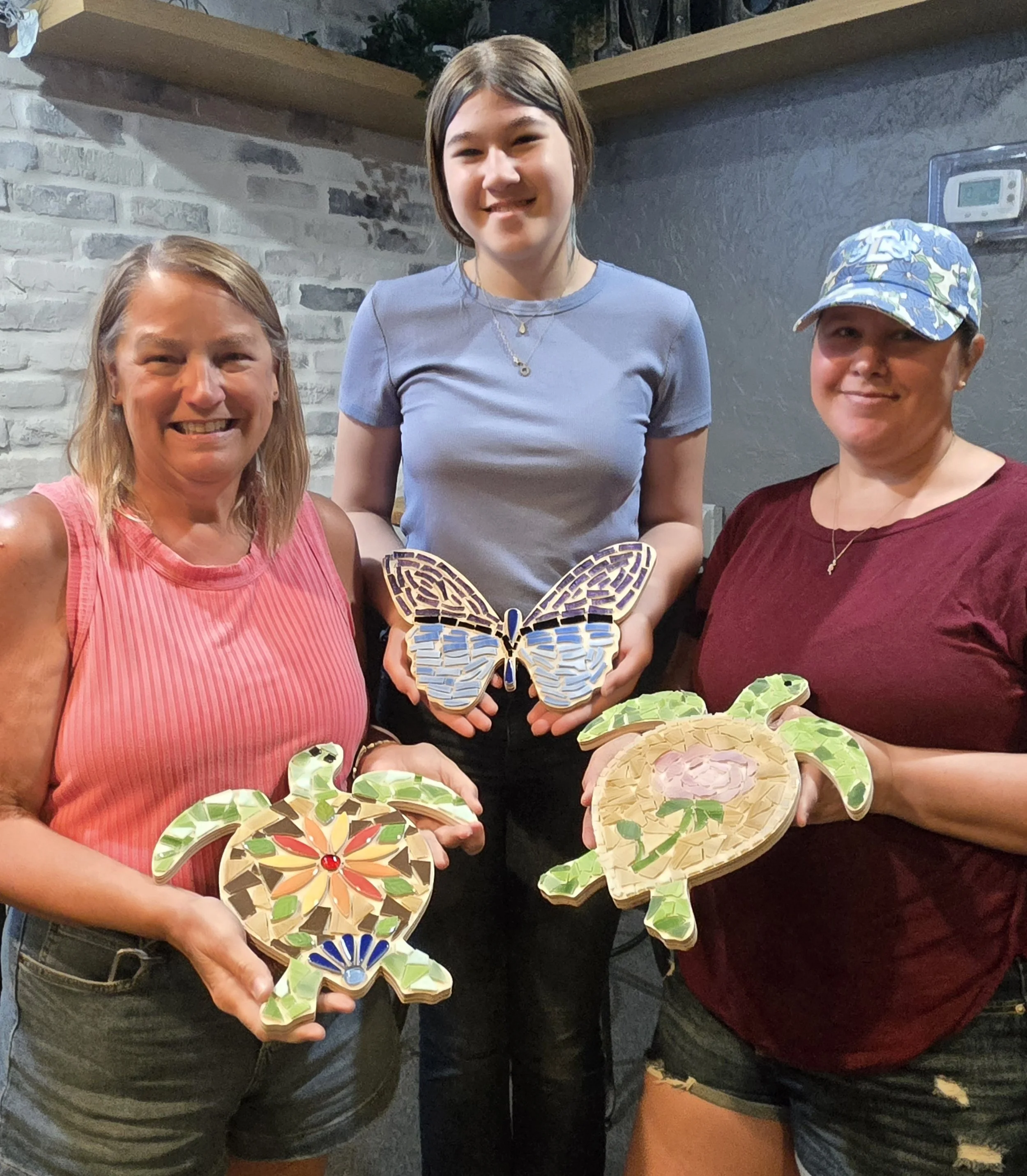 Three women holding mosaic butterfly-shaped and round art pieces in an indoor setting with a brick wall background.