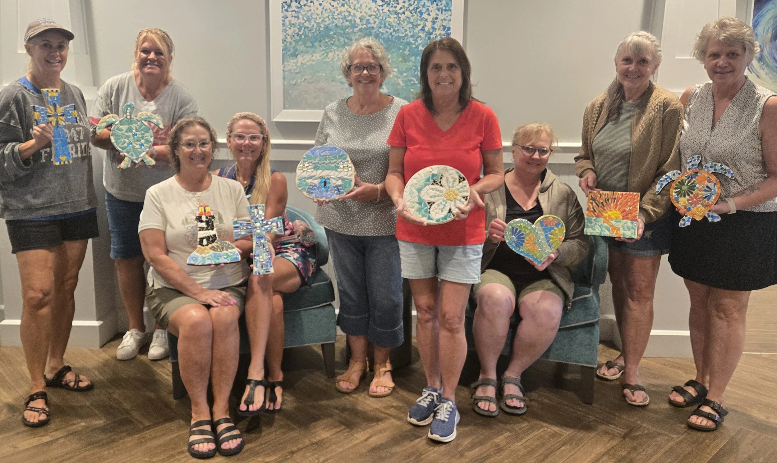 A group of ten women posing indoors, some seated and some standing, holding decorated mosaic-style art pieces, with artwork on the wall behind them.