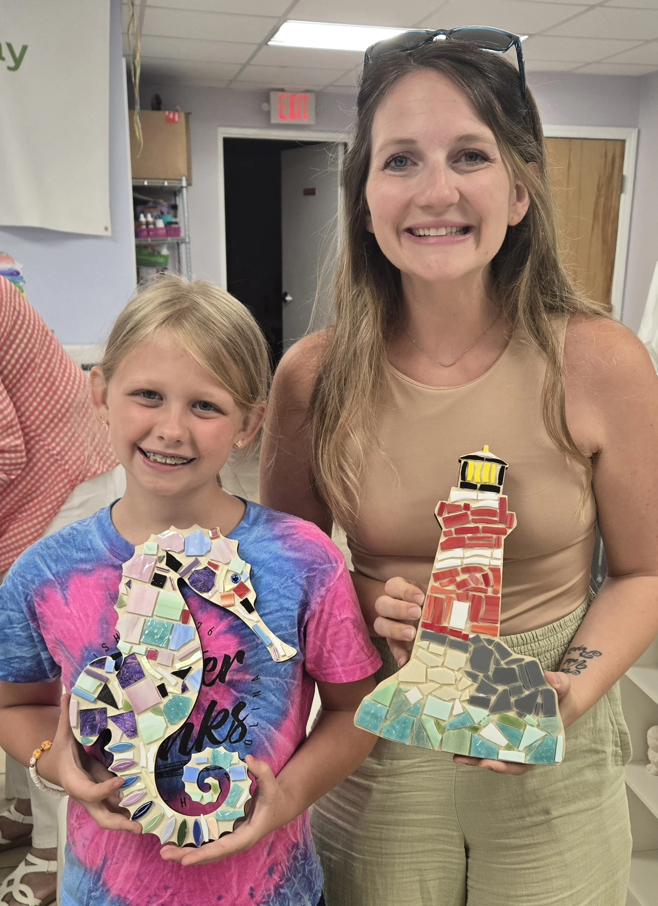 A smiling young girl holding a colorful mosaic letter G and a woman holding a mosaic I, both displaying mosaic art projects in the shape of letters.