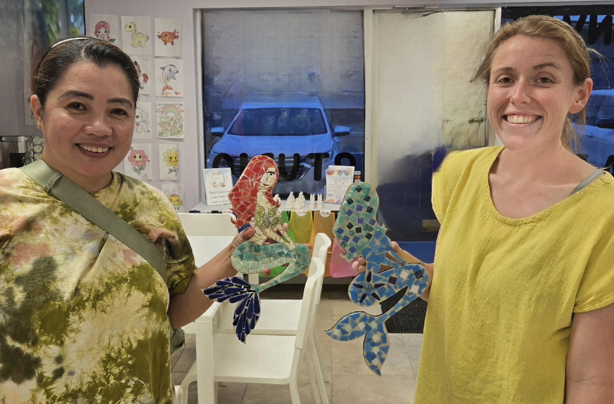 Two women smiling and holding colorful mosaic rooster and hen sculptures inside a shop, with drawings of animals on the wall behind them and a car visible through the window.