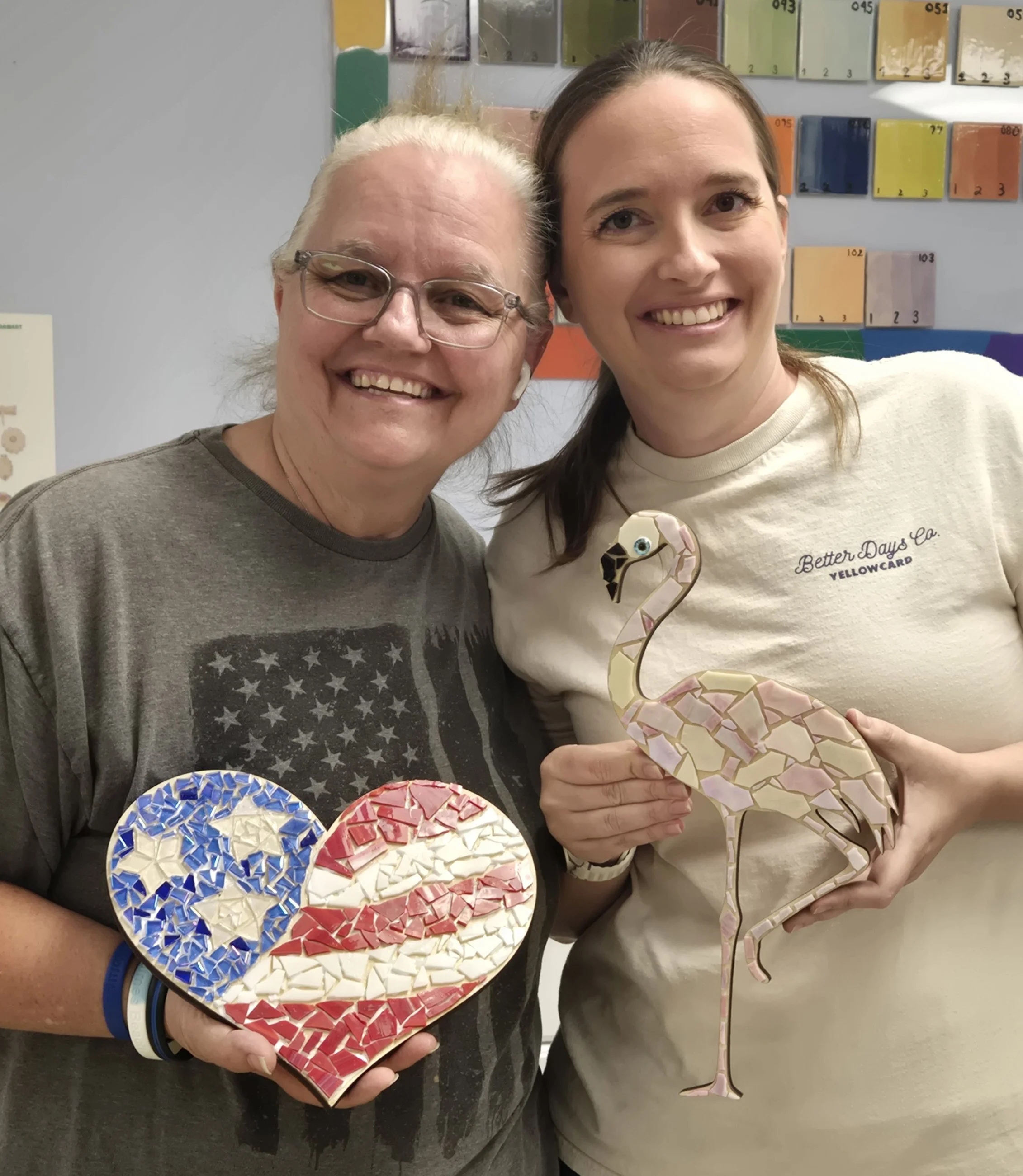 Two women smiling, holding mosaic flamingo art pieces. The woman on the left is wearing glasses and a gray shirt with a star pattern, and the woman on the right has long brown hair and is wearing a white t-shirt. Behind them is a wall with colorful c