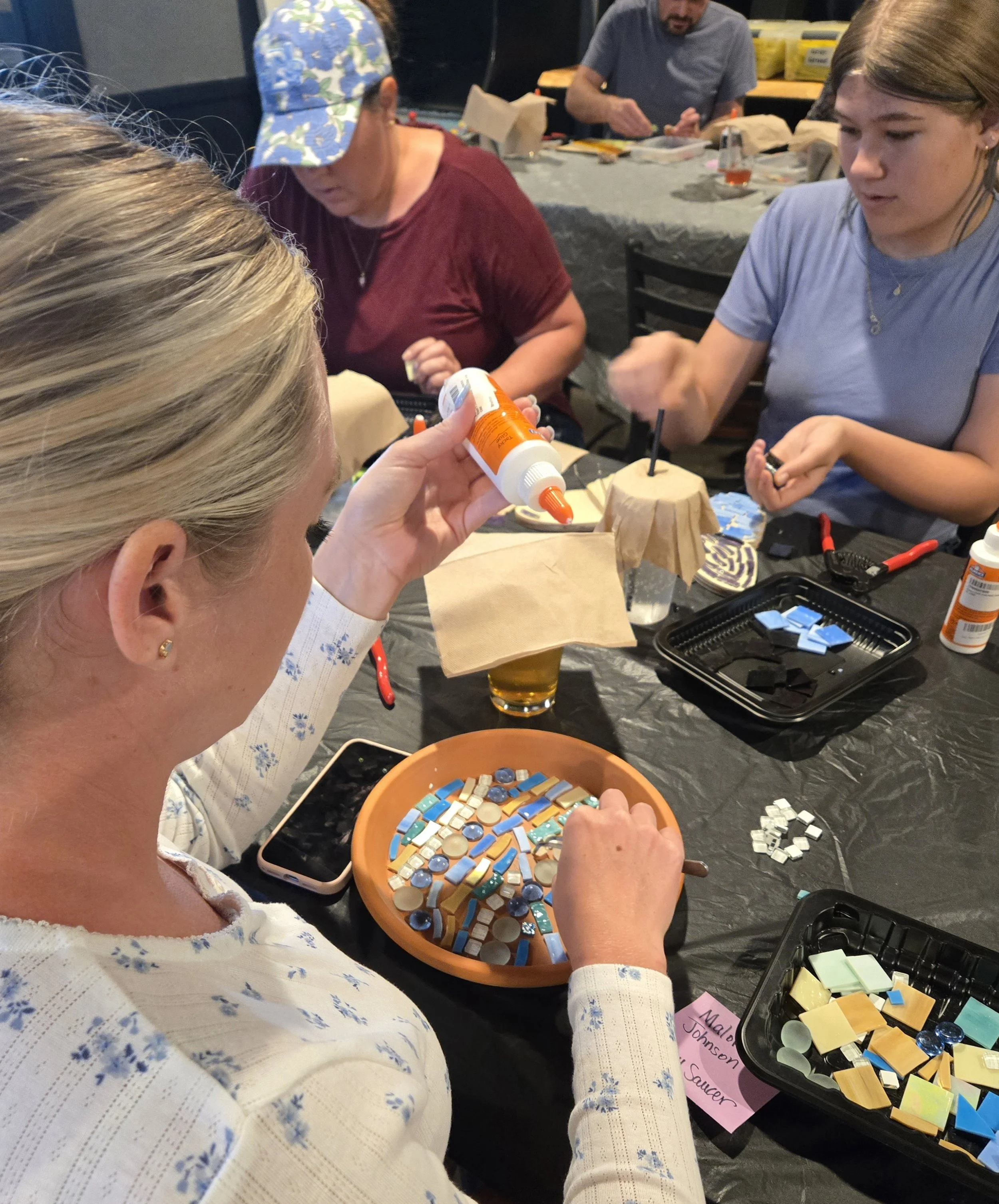 People working on a mosaic art project with tiles and glue in a craft workshop, with various supplies on the table.