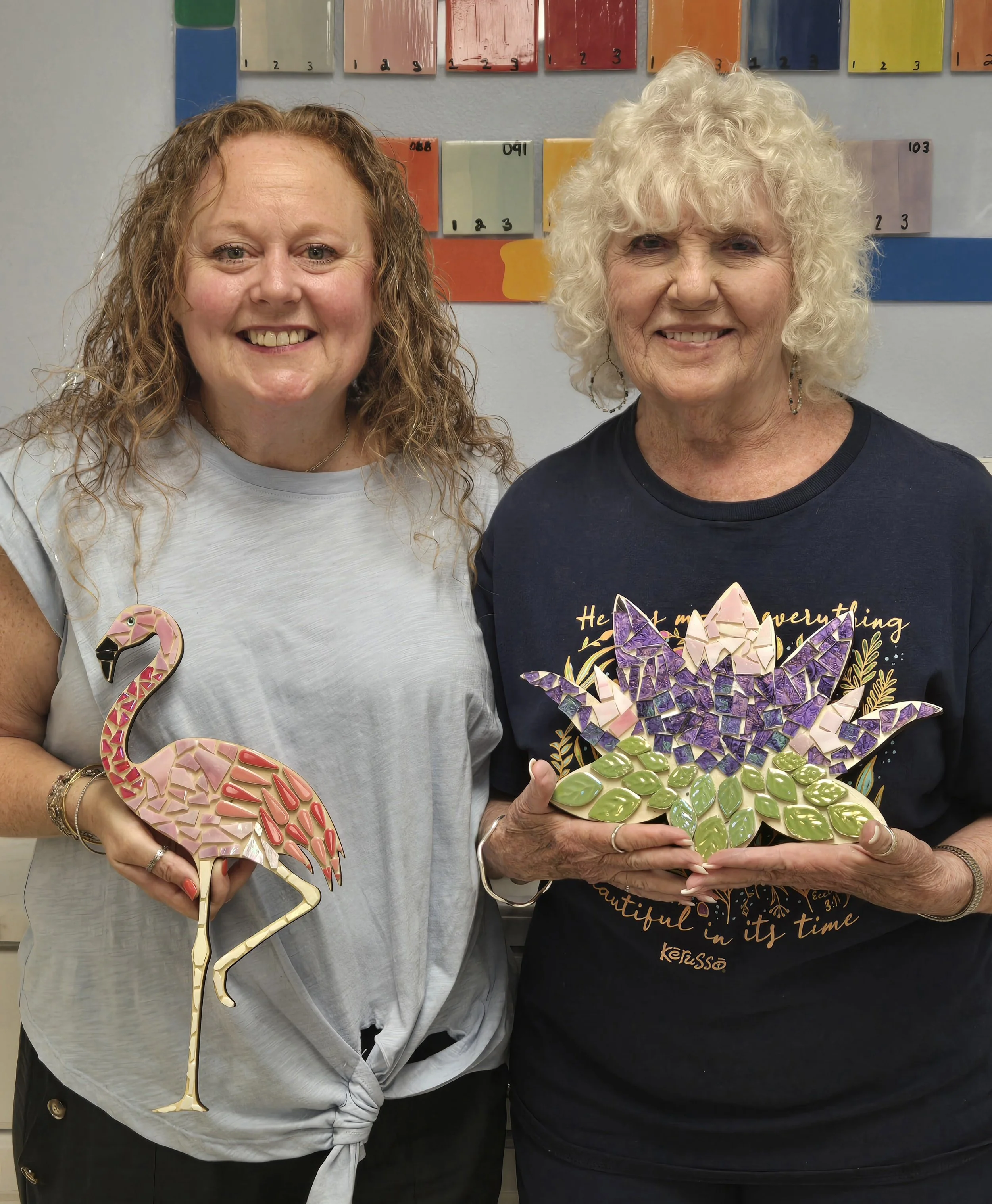 Two women smiling, standing indoors in front of color swatches on a wall. One woman holds a decorative flamingo, the other holds a colorful mosaic flower-shaped object.