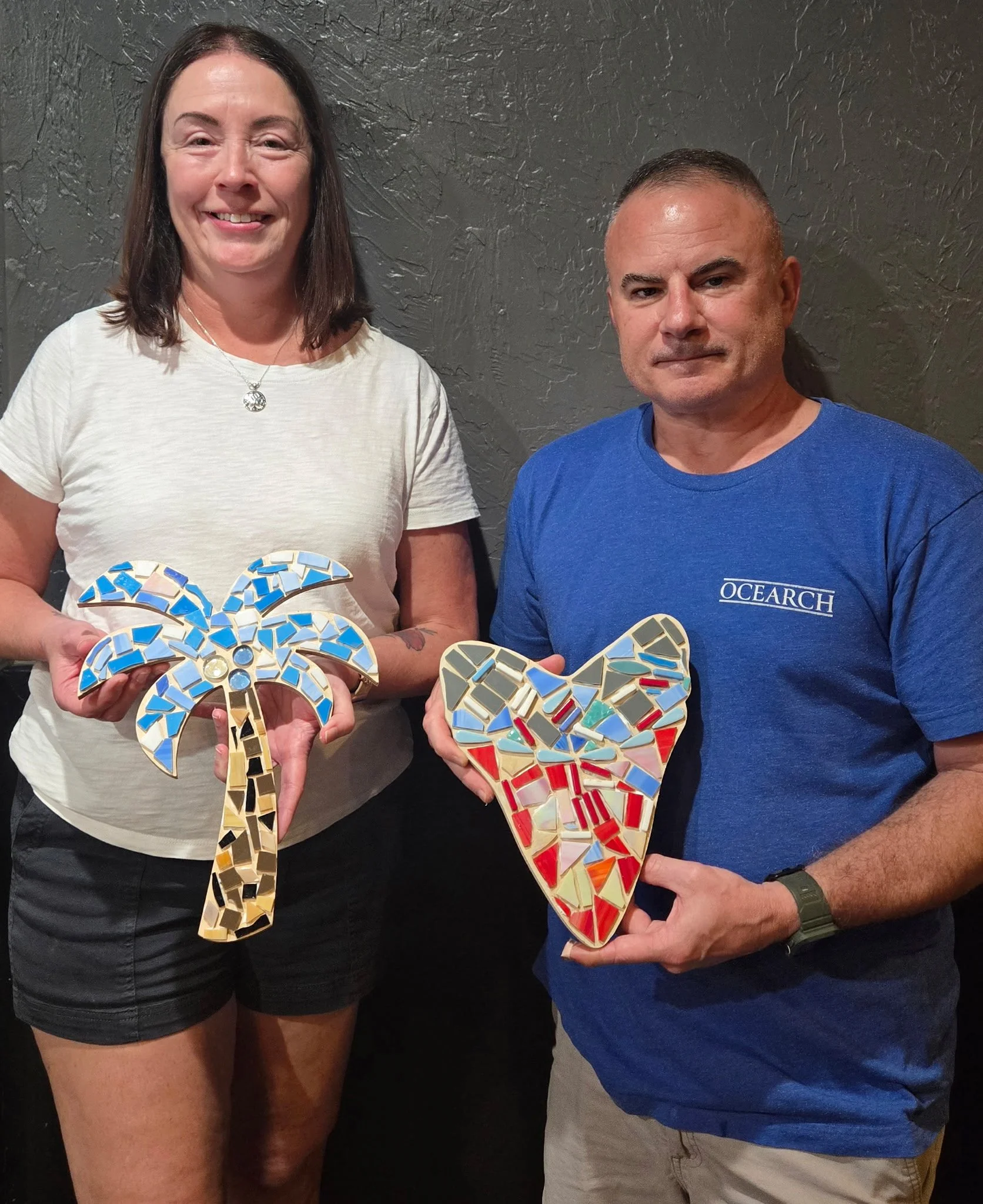 A woman and a man each holding a mosaic heart-shaped artwork. The woman is wearing a white t-shirt and black shorts, smiling, with dark hair. The man is wearing a blue t-shirt with white lettering that says 'OCEARCH' and beige shorts, with a serious 