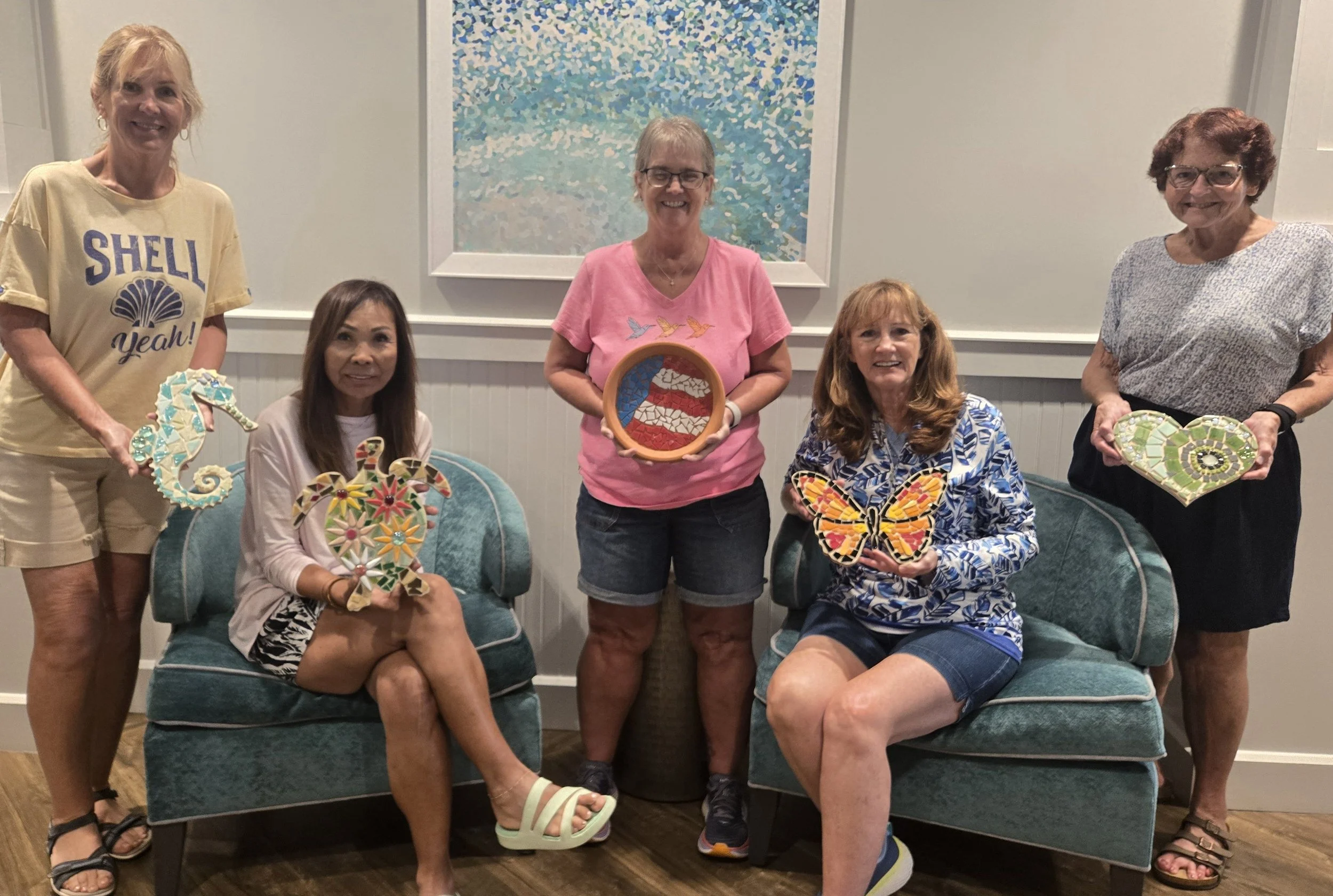Six women in a living room holding handmade mosaic art pieces, some seated on a green cushioned chair, some standing, with a large seascape painting on the wall behind them.