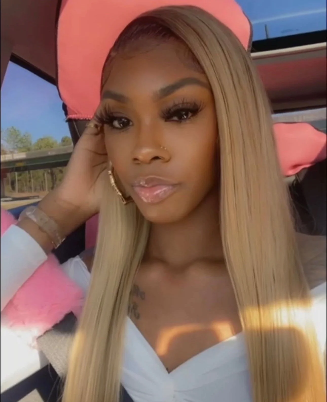 A woman with long blonde hair taking a selfie in what appears to be a vehicle, wearing makeup and large eyelashes, with a pink bow in her hair, a nose piercing, and small tattoos on her chest.