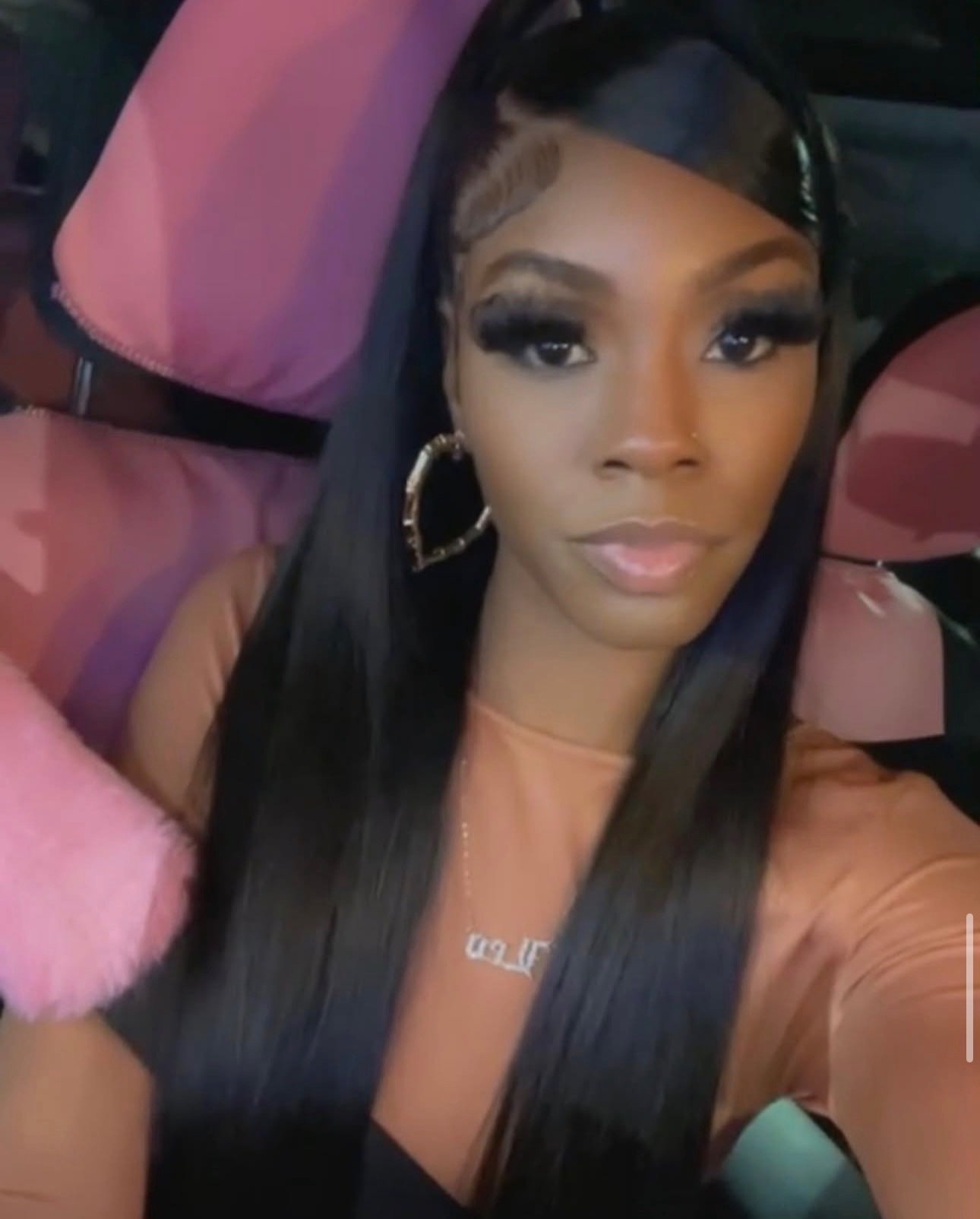 A woman with long black hair, hoop earrings, and a nose piercing taking a selfie in a vehicle with pink and purple cushioned seats.