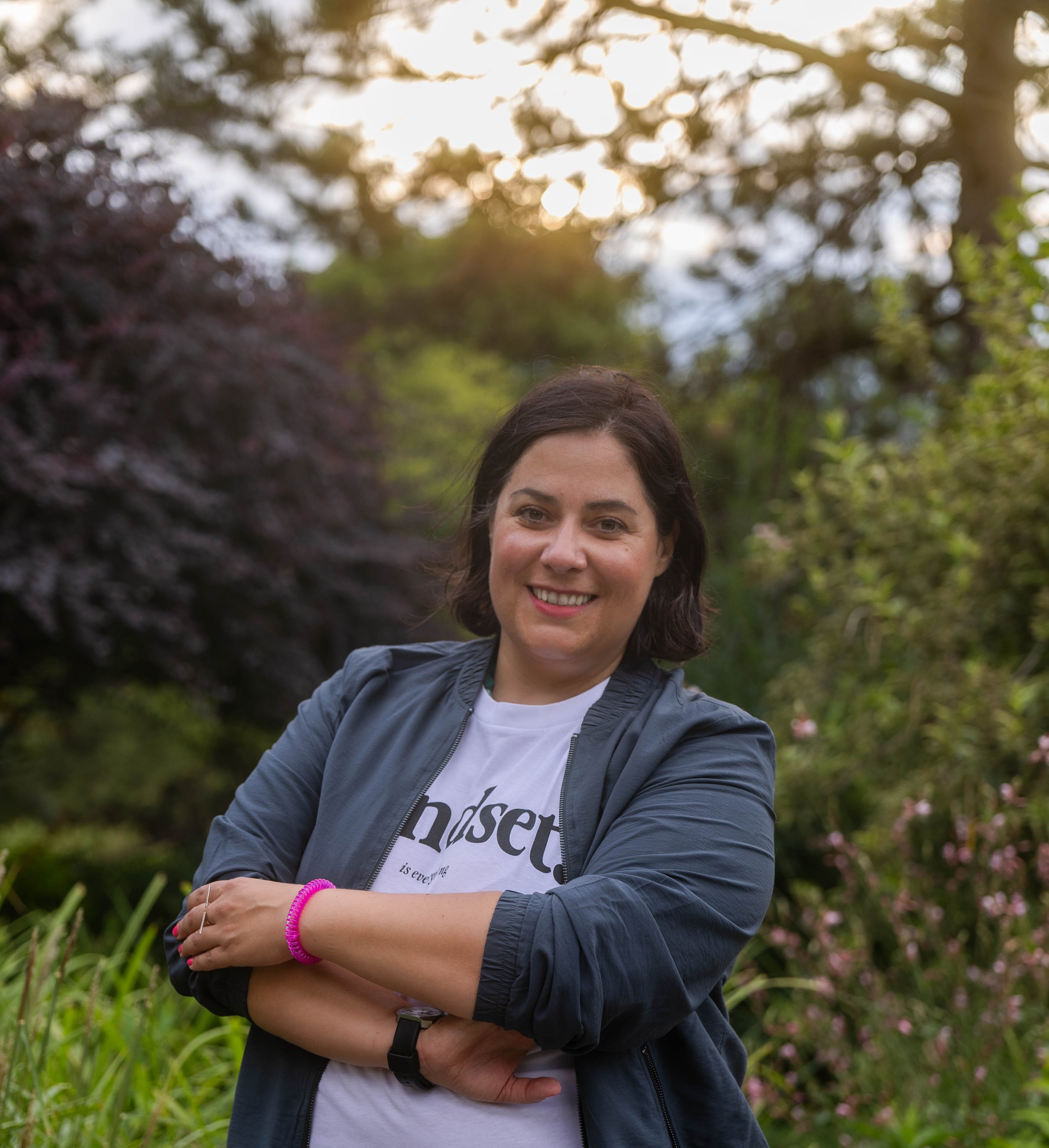 Angelika Fabian, eine lächelnde Frau mit dunklen Haaren posiert in einem Garten bei Sonnenuntergang, umgeben von grünen Pflanzen und Bäumen.