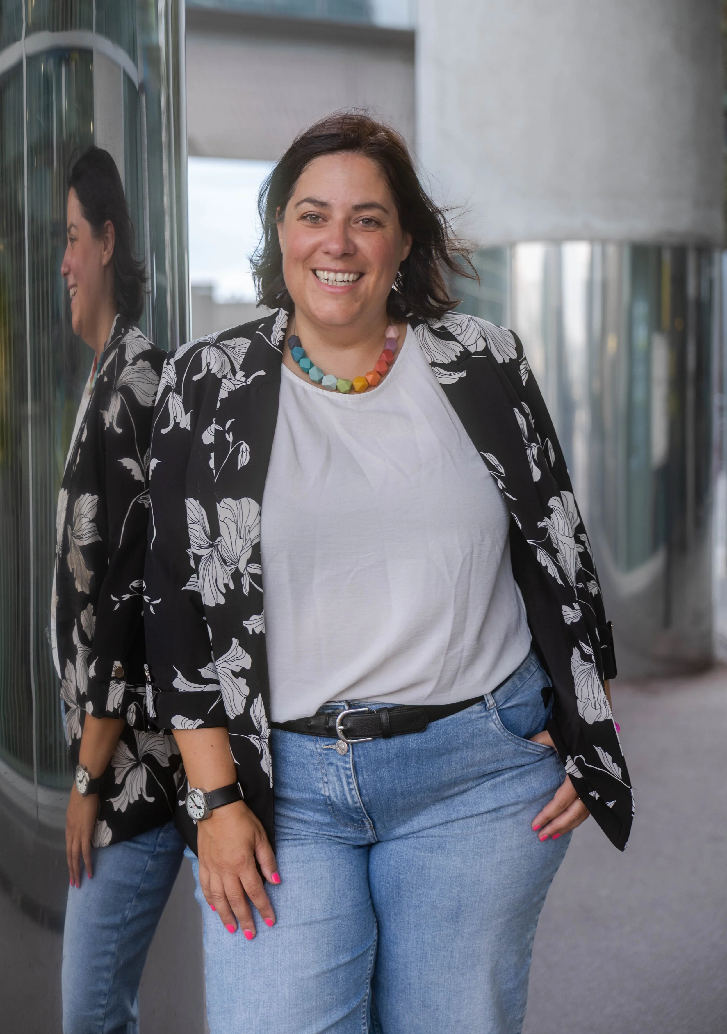 Angelika Fabian, eine lachende Frau trägt eine schwarze Jacke mit weißen Blumen, eine weiße T-Shirt und blaue Jeans, steht vor einem modernen Gebäudeteil mit Glas und Beton, trägt bunte Halskette, Armband, Uhren, pinkfarbene Fingernägel.