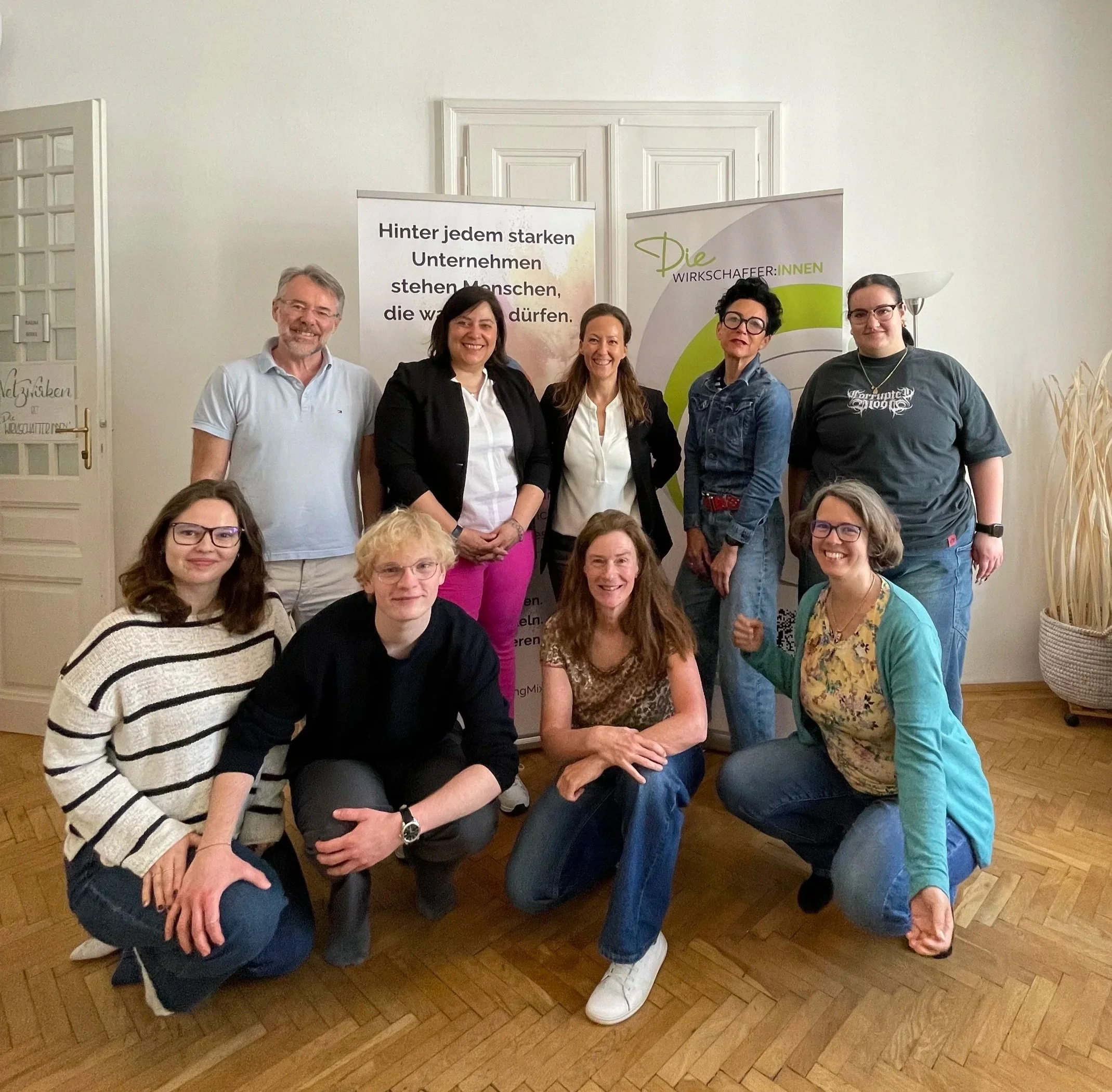 Fünf Frauen und ein Mann posieren in einem Raum, stehen im Vordergrund, einige knien, vor Bannern mit Texten, einer wand, einem Sofa und Pflanzen.