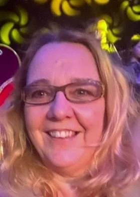 Nahaufnahme einer lachenden Frau mit Brille und lockigem, blondem Haar bei einer Feier im Neonlicht.