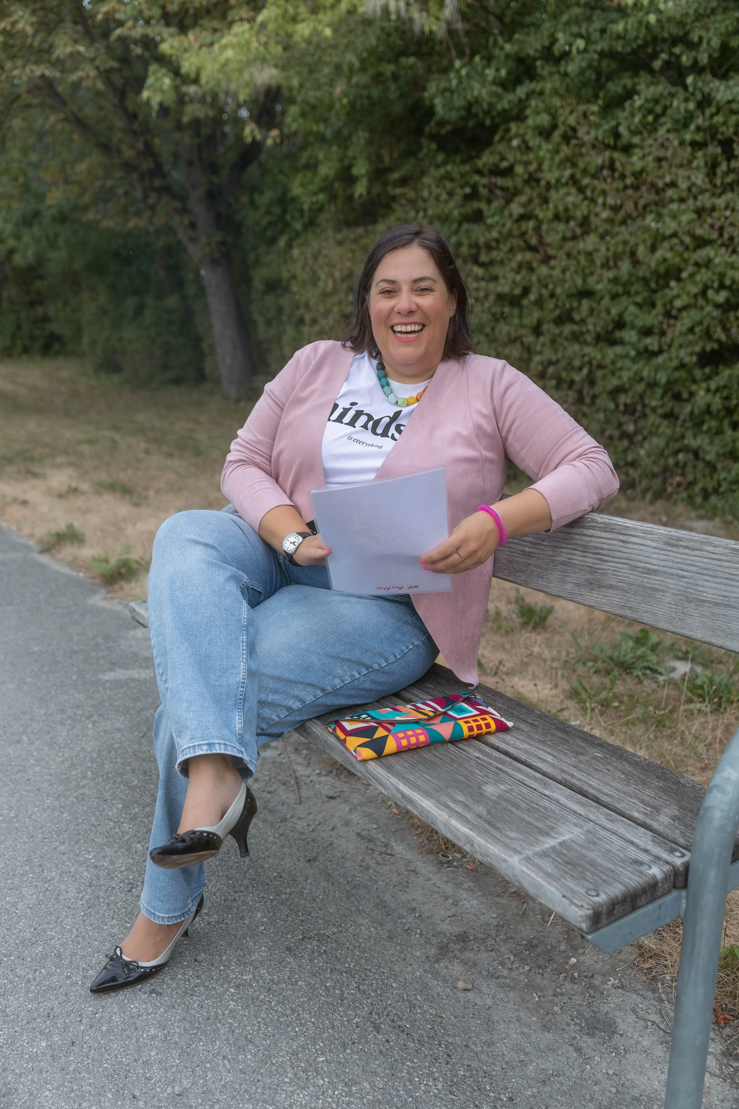 Angelika Fabian, eine lachende Frau sitzt auf einer Parkbank im Freien, trägt eine pinke Jacke, eine T-Shirt mit Text, blaue Jeans, schwarze High Heels, und hält Papiere in der Hand. Vor der Bank liegt eine bunte Stofftasche.
