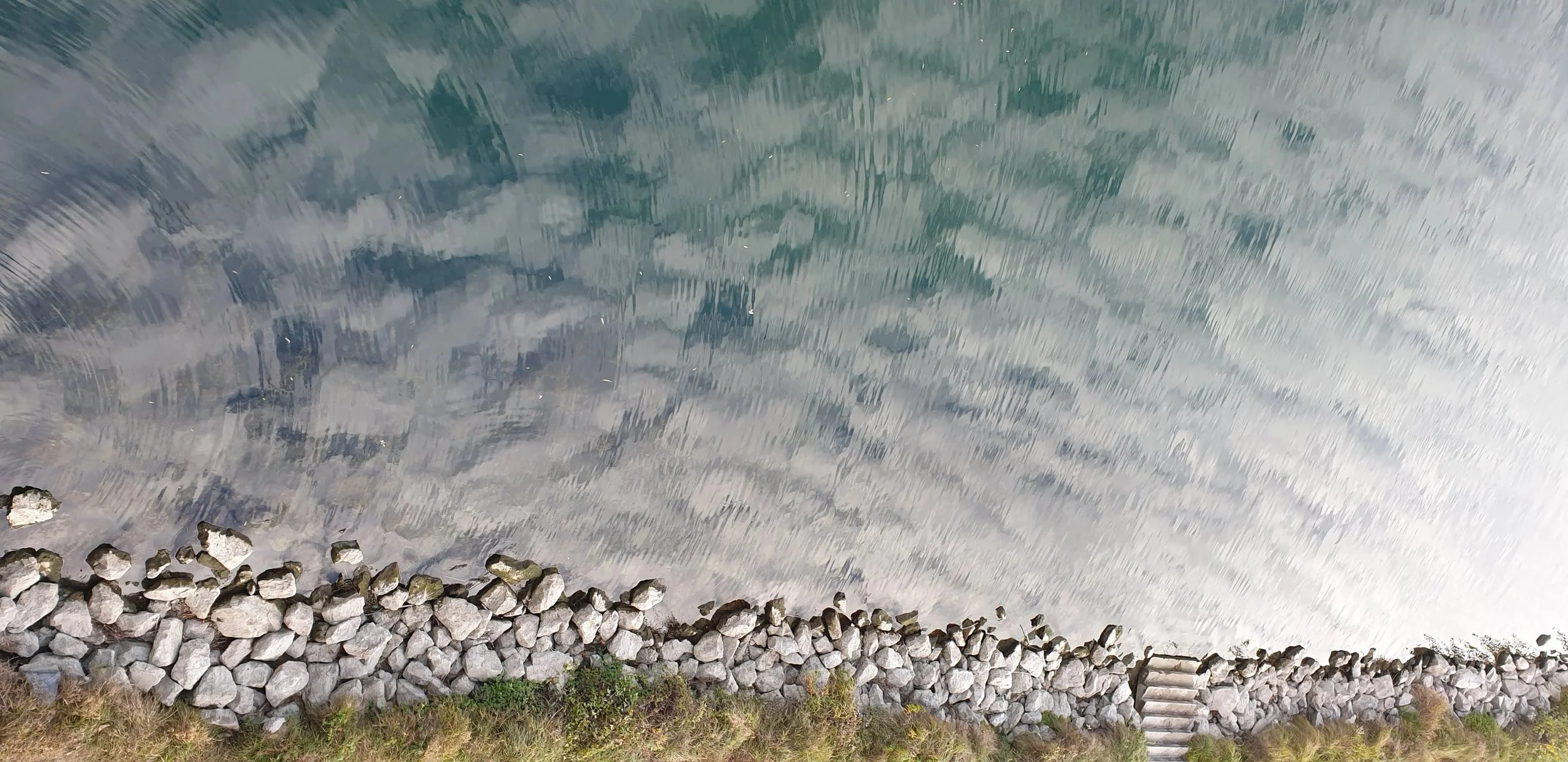 Steinpfad entlang eines Sees mit grün-blauem Wasser, Steine am Ufer und ein Holzsteg.