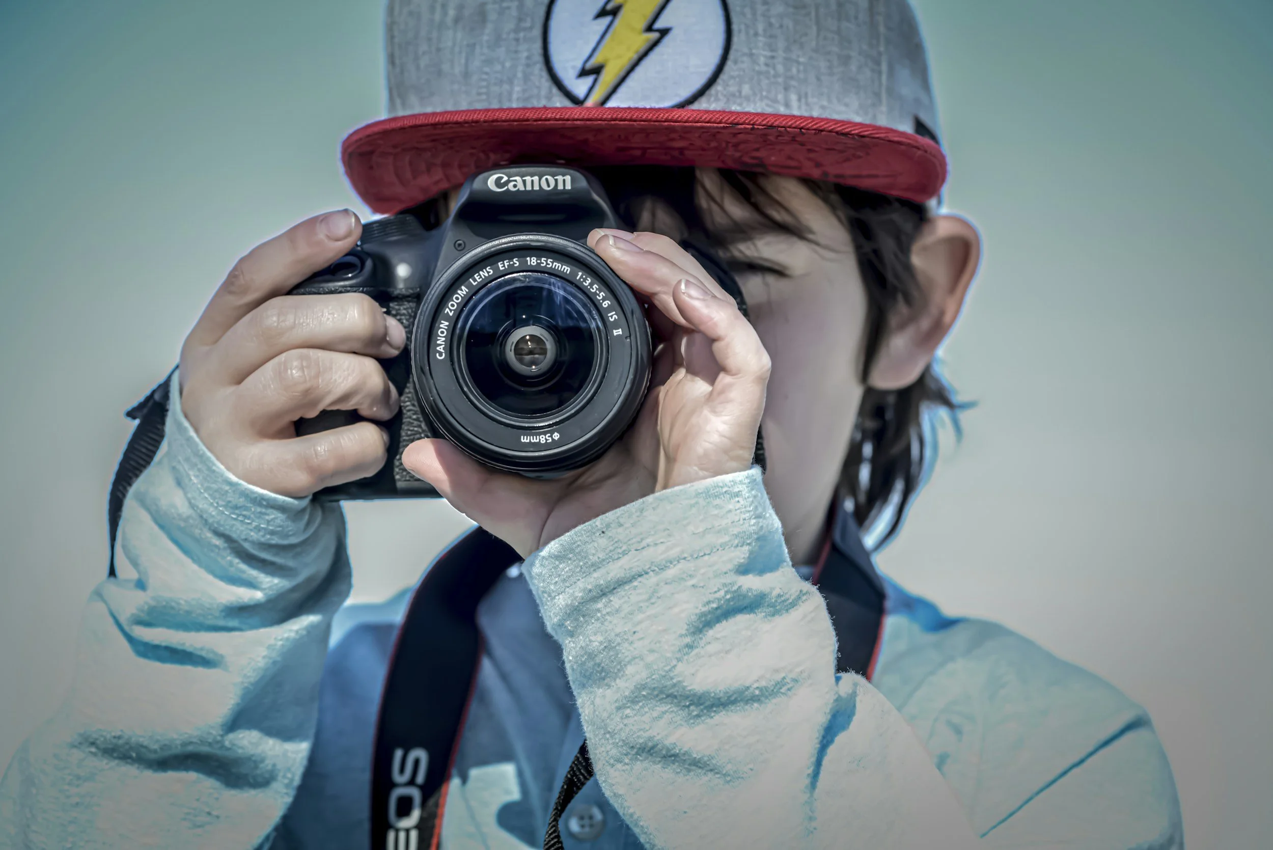 Une personne tenant un appareil photo Canon, portant un bonnet gris avec un logo d'éclair, une veste gris clair et une écharpe bleu