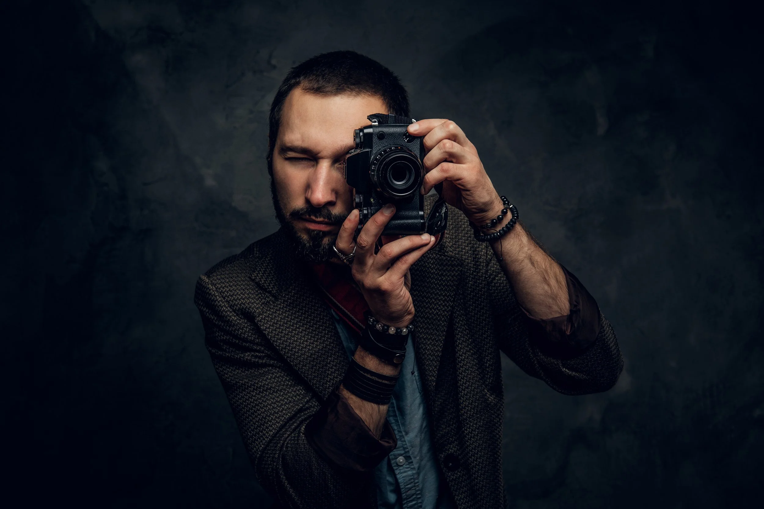 Homme avec une barbe prenant une photo avec un appareil photo contre un fond sombre.