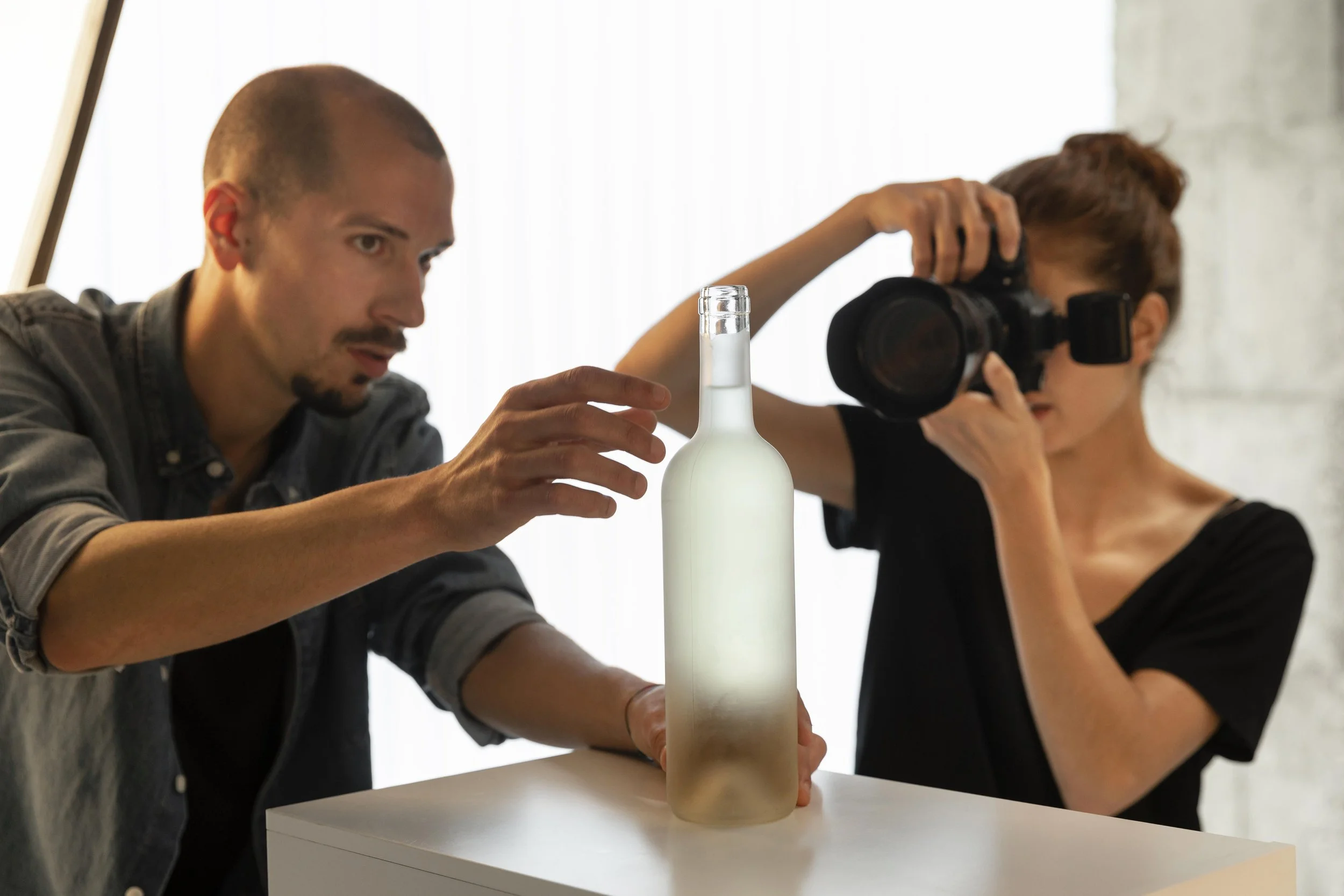 Deux personnes prennent une photo d'une bouteille opaque sur une table, une avec un appareil photo et l'autre en manipulant la bouteille.