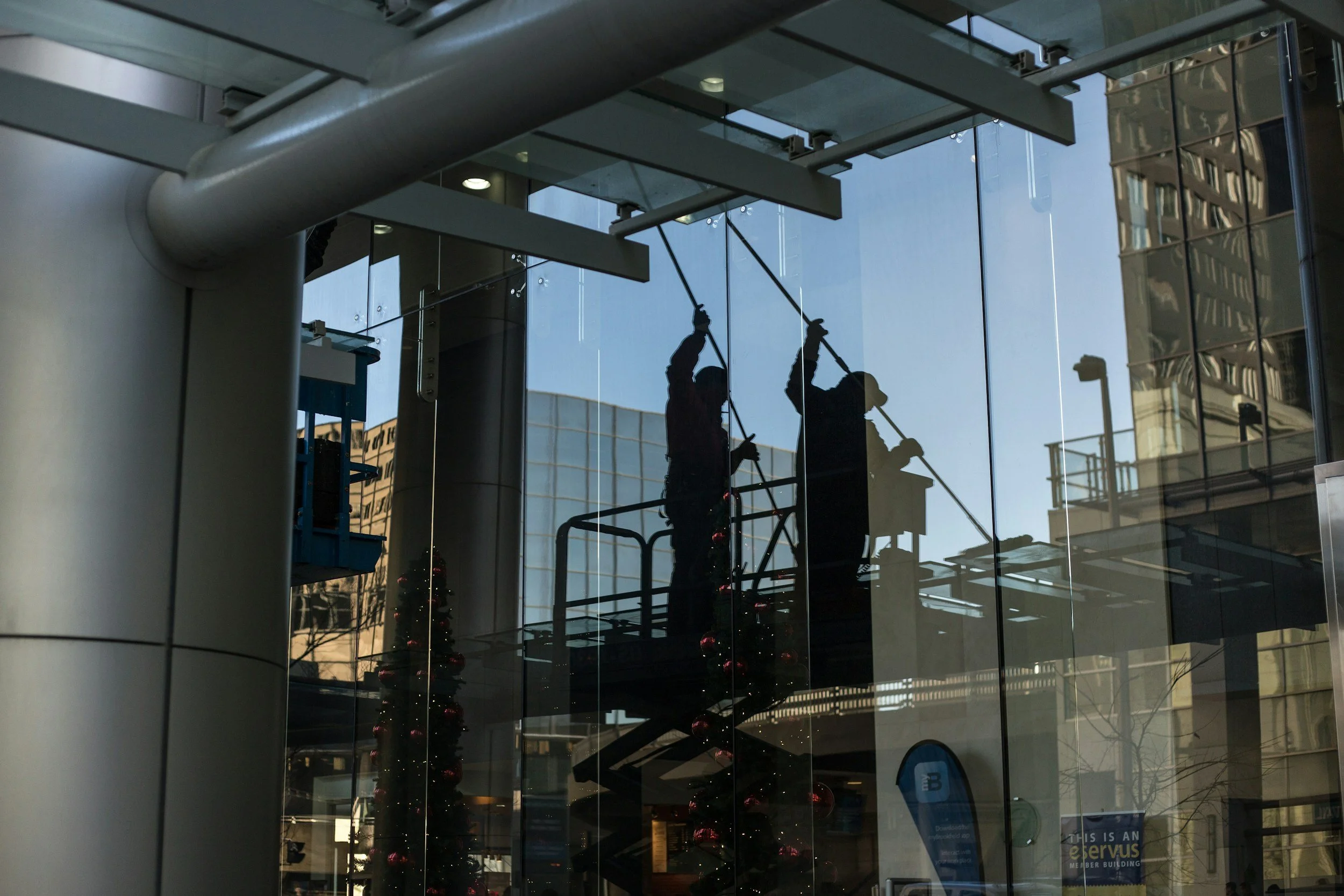 Reflected image of two workers cleaning windows on a high-rise building, with Christmas trees and city buildings visible inside.