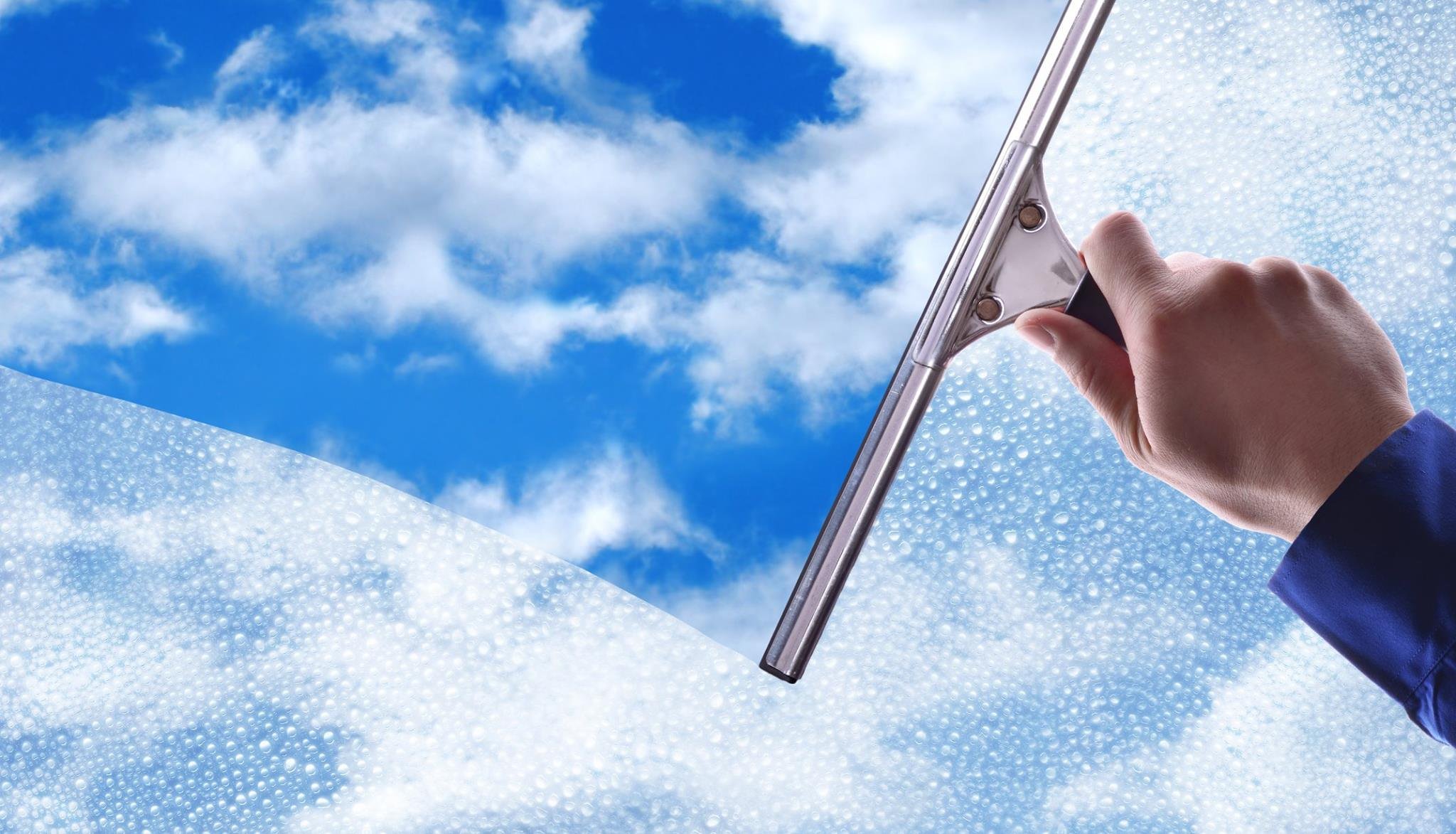A person cleaning a glass window with a squeegee, revealing a blue sky with white clouds behind the glass.