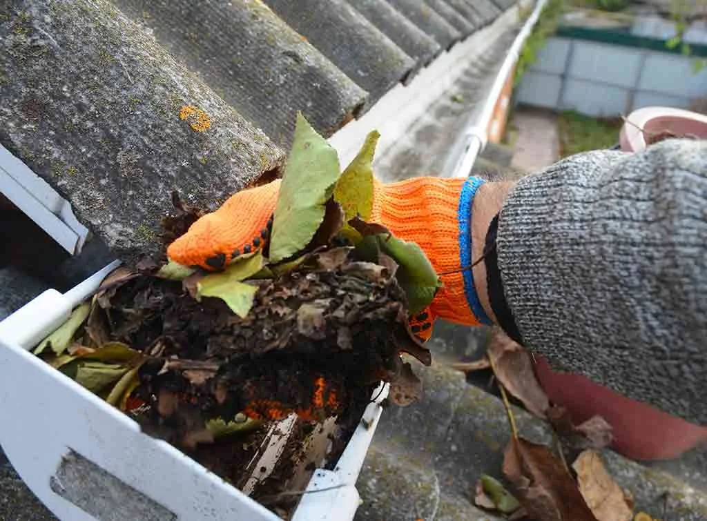 Person wearing orange gloves and a gray sweater raking leaves on a roof under cloudy sky.