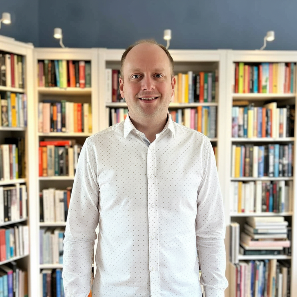 Auteur in een witte, gedessineerde blouse in een bibliotheek met witte boekenplanken en veel boeken.