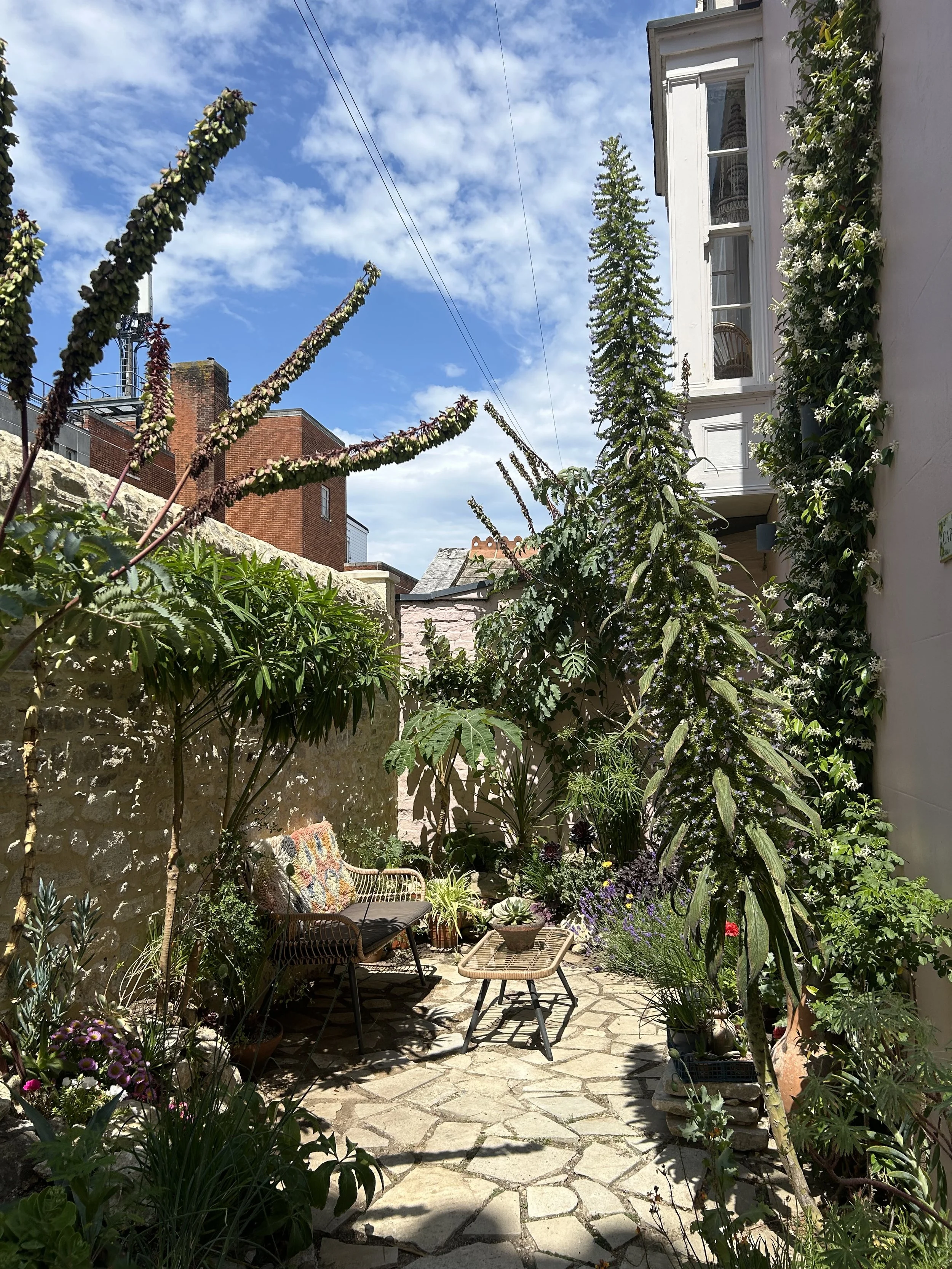 A cozy outdoor garden patio with a stone pathway, surrounded by lush green plants and flowers, a wicker bench with colorful cushions, a small table with a potted plant, and a stone wall with a white building with bay windows on the right. The sky is partly cloudy and sunny.