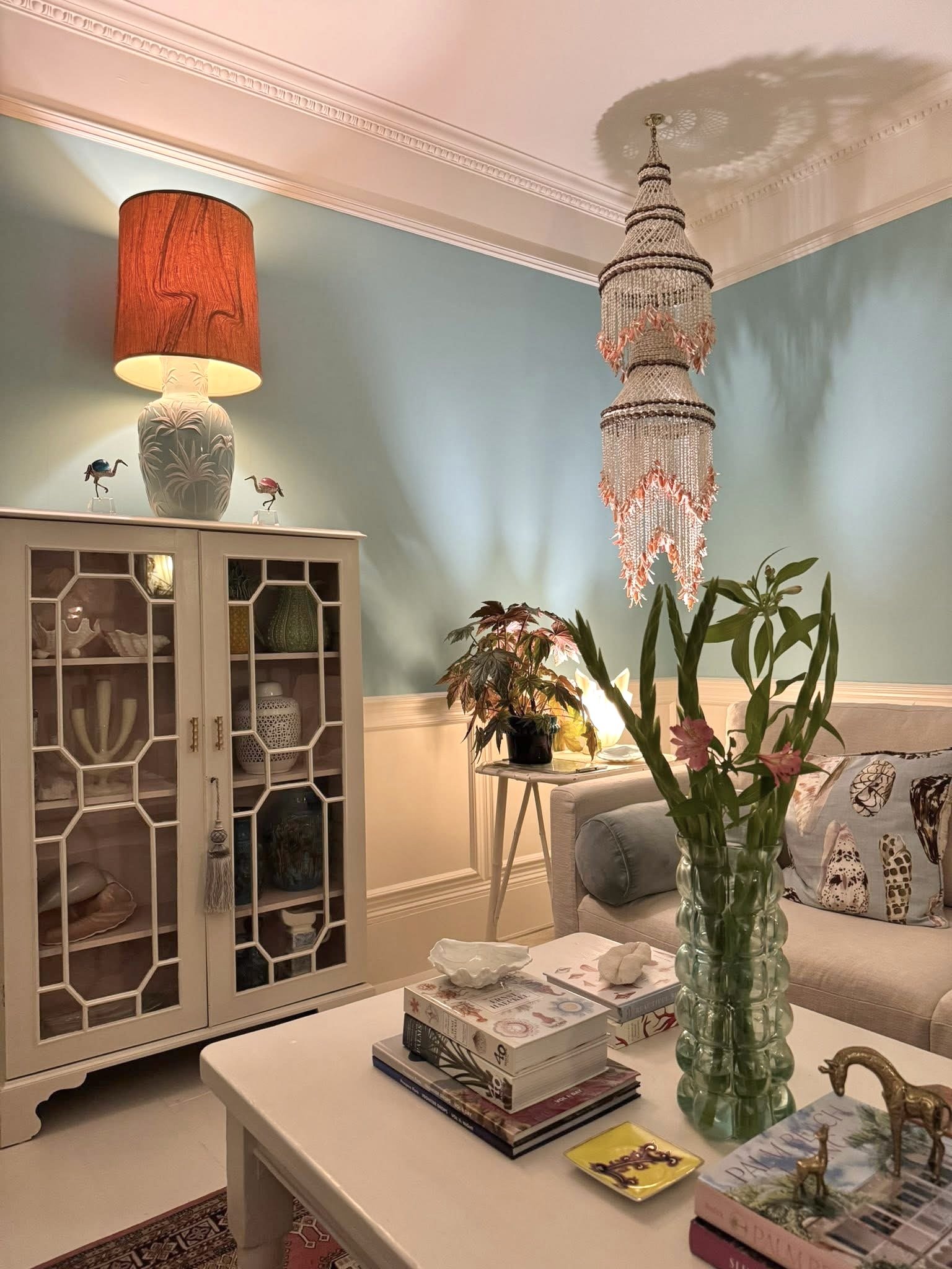 A cozy living room with a white coffee table holding a stack of books and a large green glass vase with pink flowers. In the background, a beige sofa with animal print pillows, a side table with a plant and a lamp, a white display cabinet with decorative items, a colorful chandelier hanging from the ceiling, and a lamp with a red shade atop the cabinet, all against a mint green wall with white crown molding.