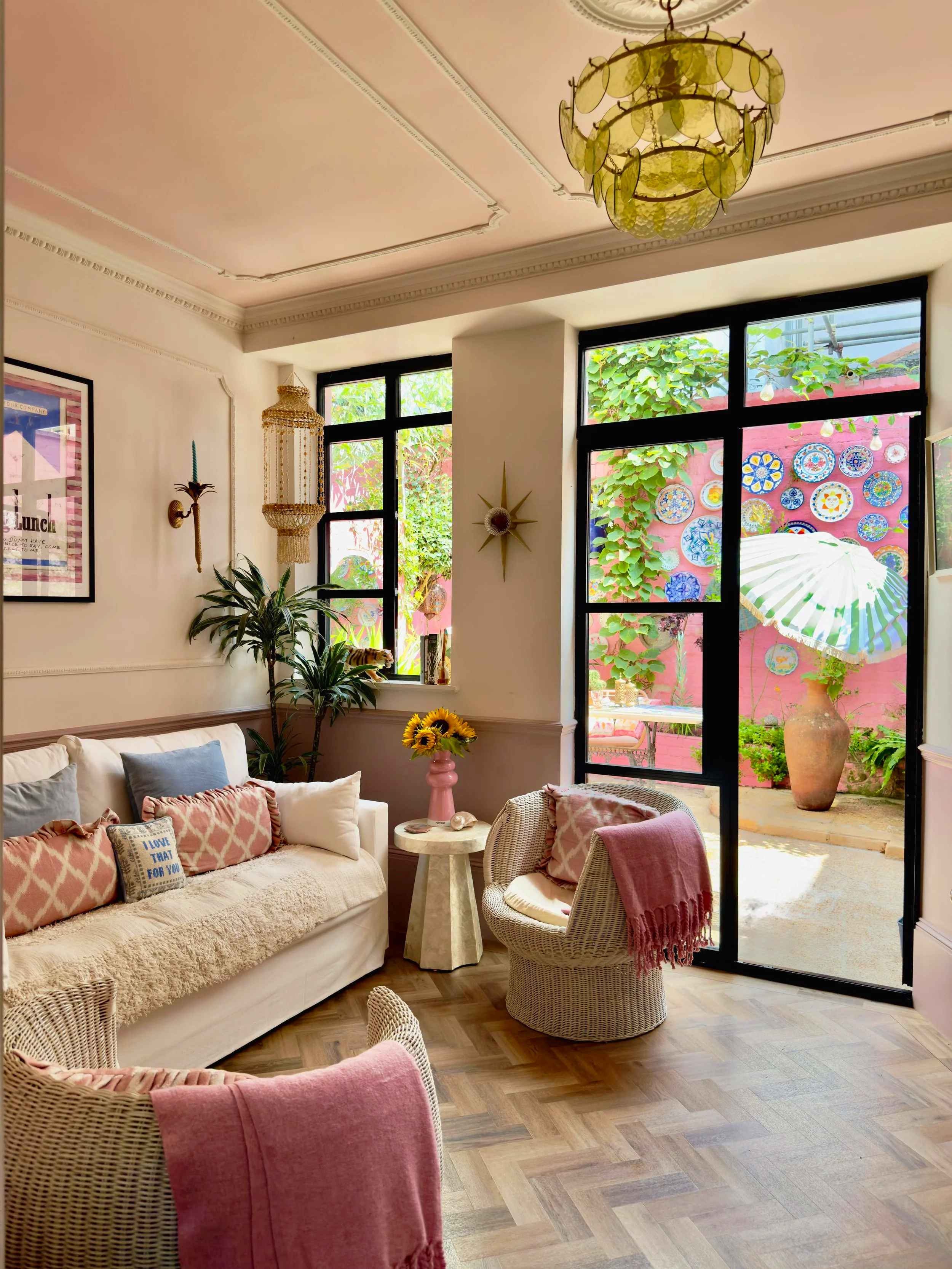 Cozy living room with large windows, cream sofa with pink and blue pillows, wicker chairs with pink blankets, a small round table with a pink vase and sunflowers, indoor plants, and an outdoor patio with colorful plates on a pink wall and a large umbrella.