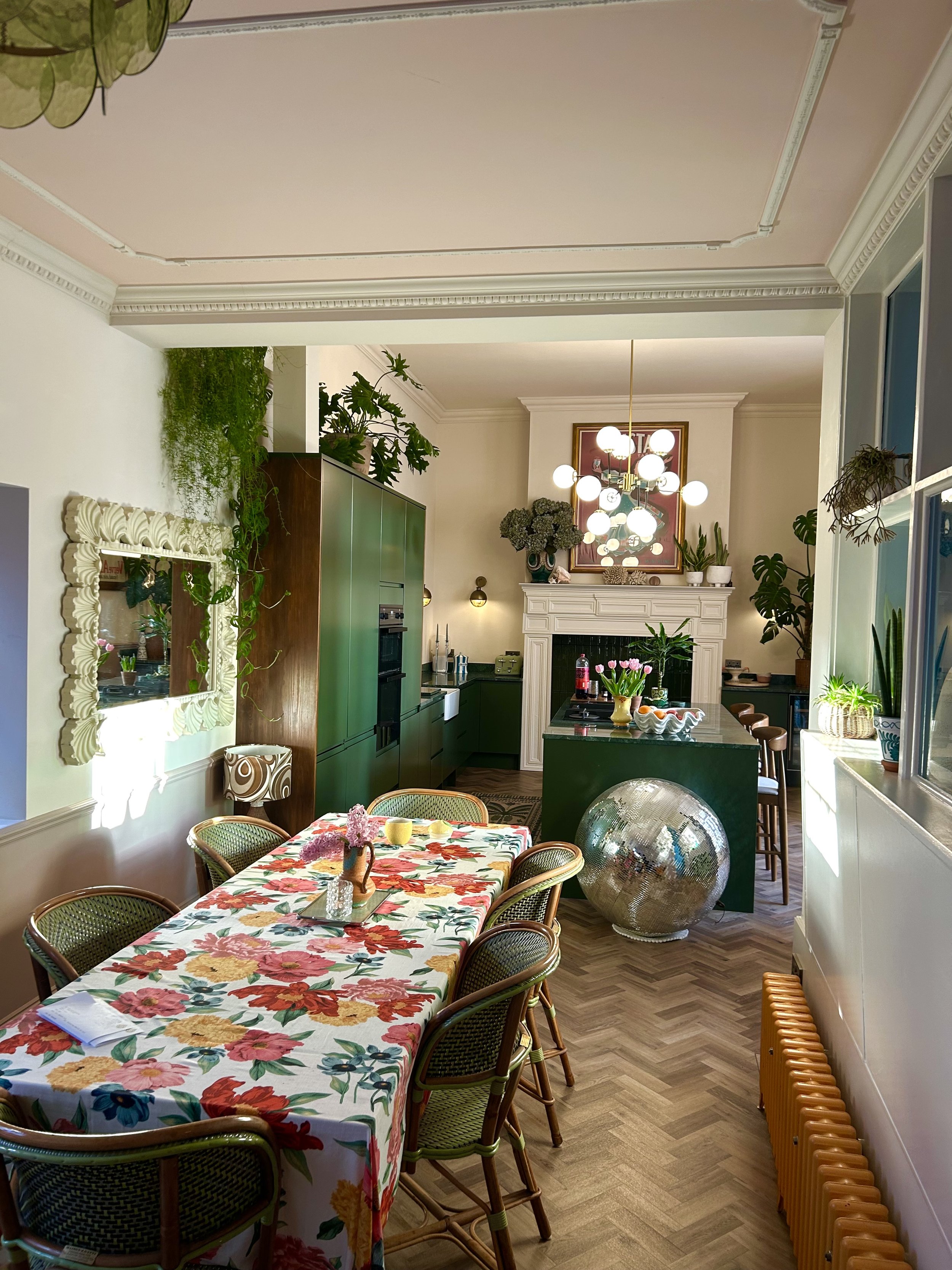 A dining room with a floral tablecloth, around 8 rattan chairs, green kitchen cabinets, a disc mirror, a fireplace with plants and a chandelier, and decorative elements including a large ball and lamps.