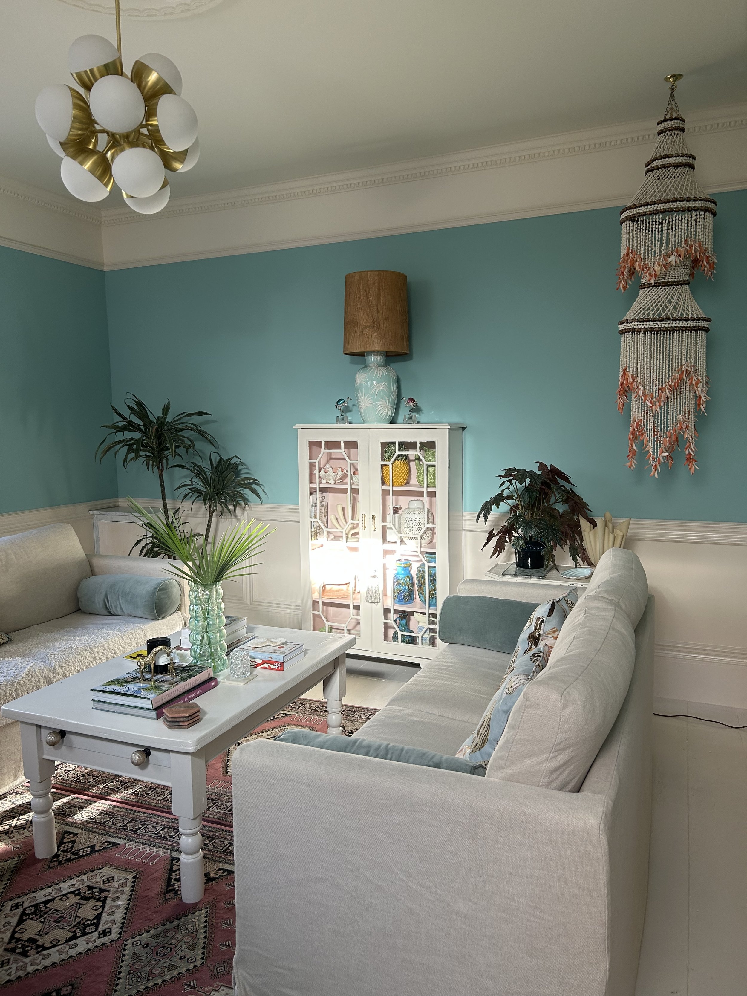 Living room with teal walls, white furniture, a white coffee table with books, plants, and decorative items, a white display cabinet, a vintage rug, and a sculptural chandelier with spherical lights.