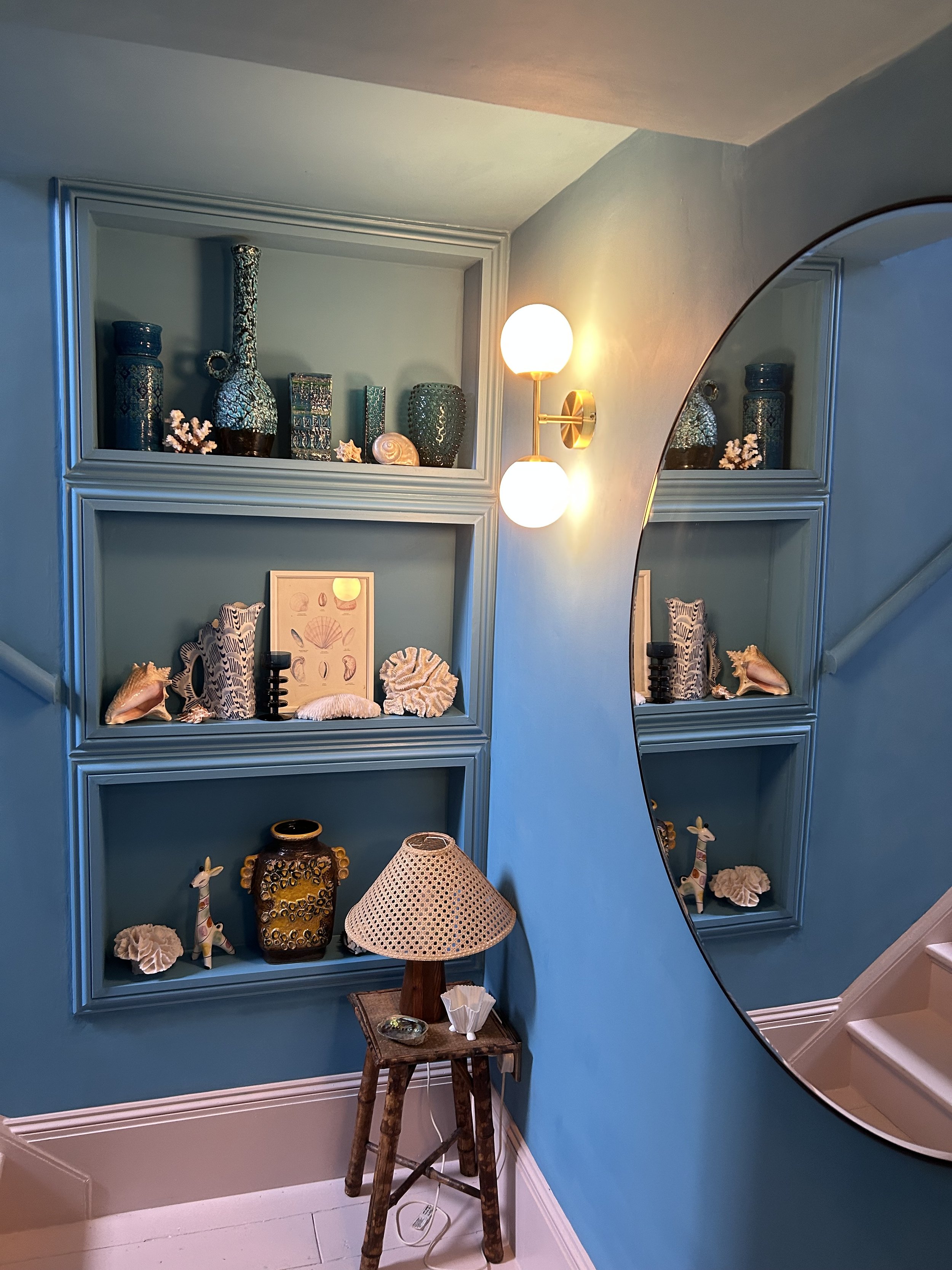 Decorative display with shelves on blue wall, filled with vases, shells, coral, and animal figurines. A round mirror is mounted on the wall, and a small wooden side table with a woven lampshade and a lamp is in front. A wall-mounted light fixture with two globes illuminates the scene.