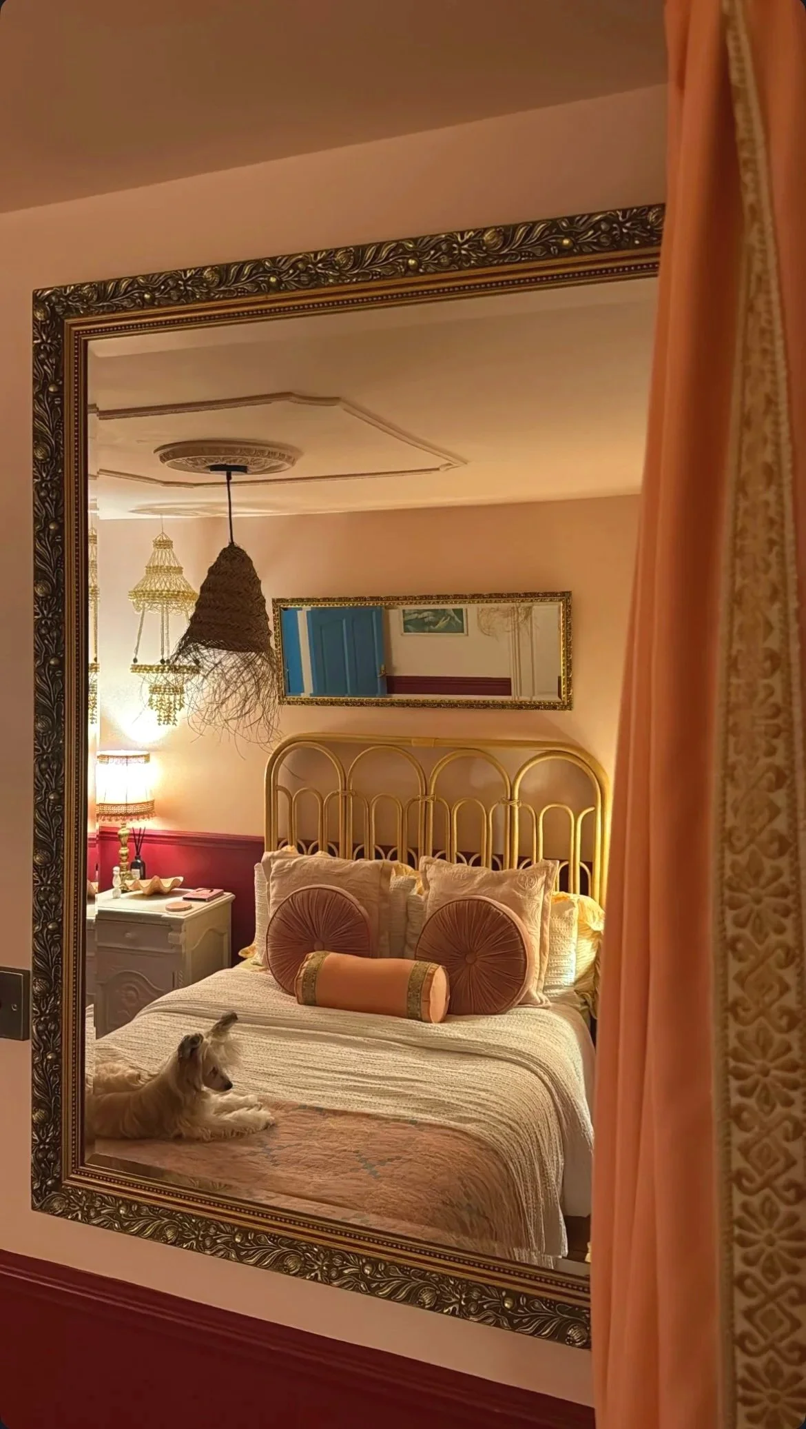 A cozy bedroom with a golden headboard, decorative pillows, a white nightstand with a lamp, and a small dog laying on the bed. The room has warm lighting, an intricate ceiling design, and a mirror reflecting a blue door.
